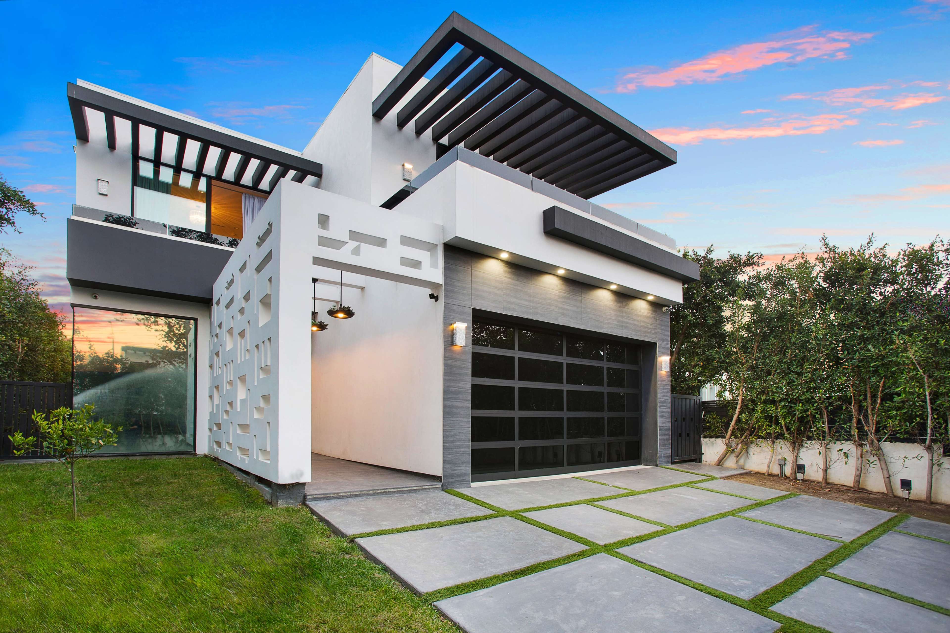 The image shows a modern house with a distinctive facade, featuring a large garage, geometric designs, and a landscaped yard.