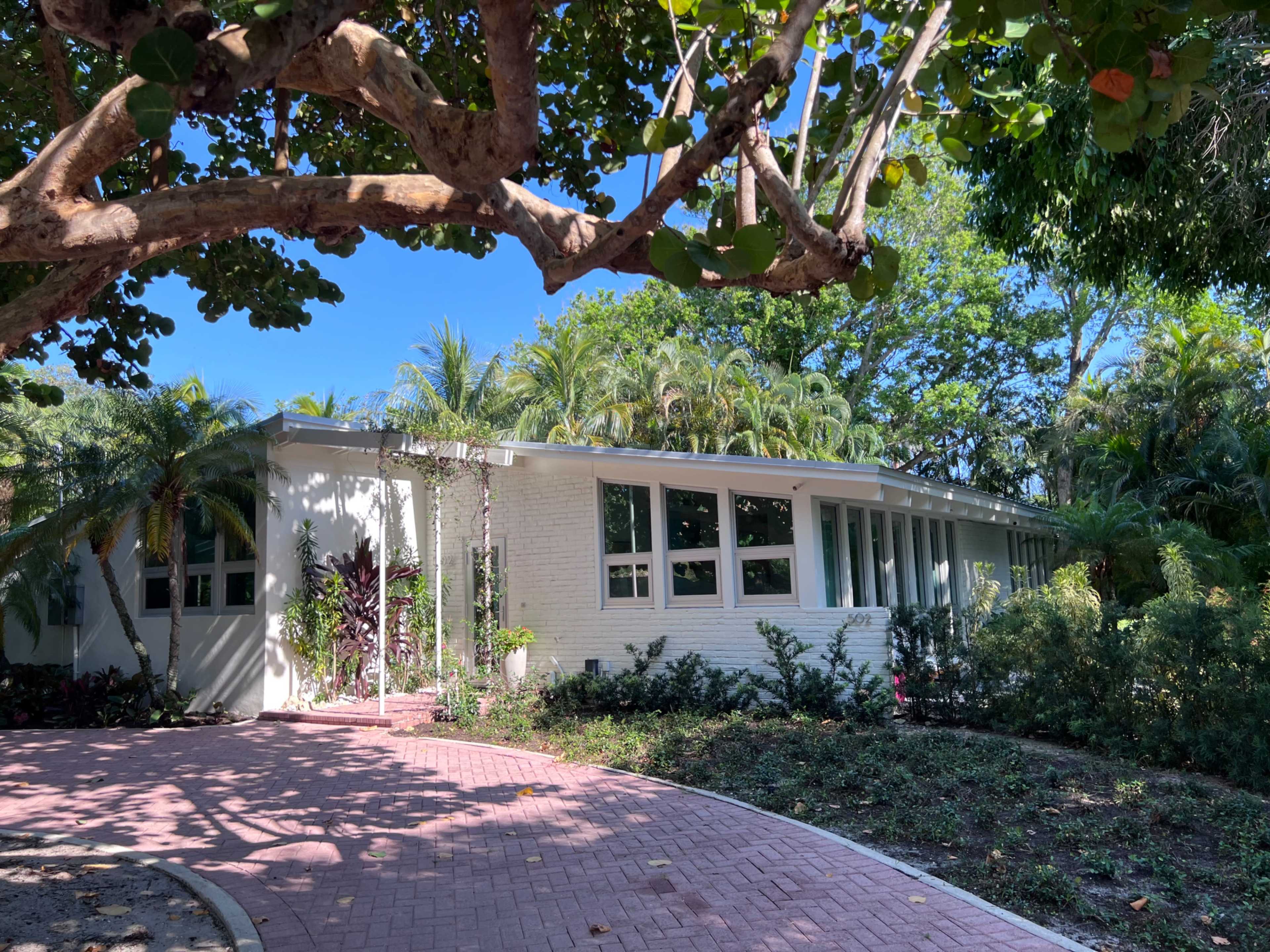 A modern white house with large windows is surrounded by tropical foliage and a winding brick pathway.