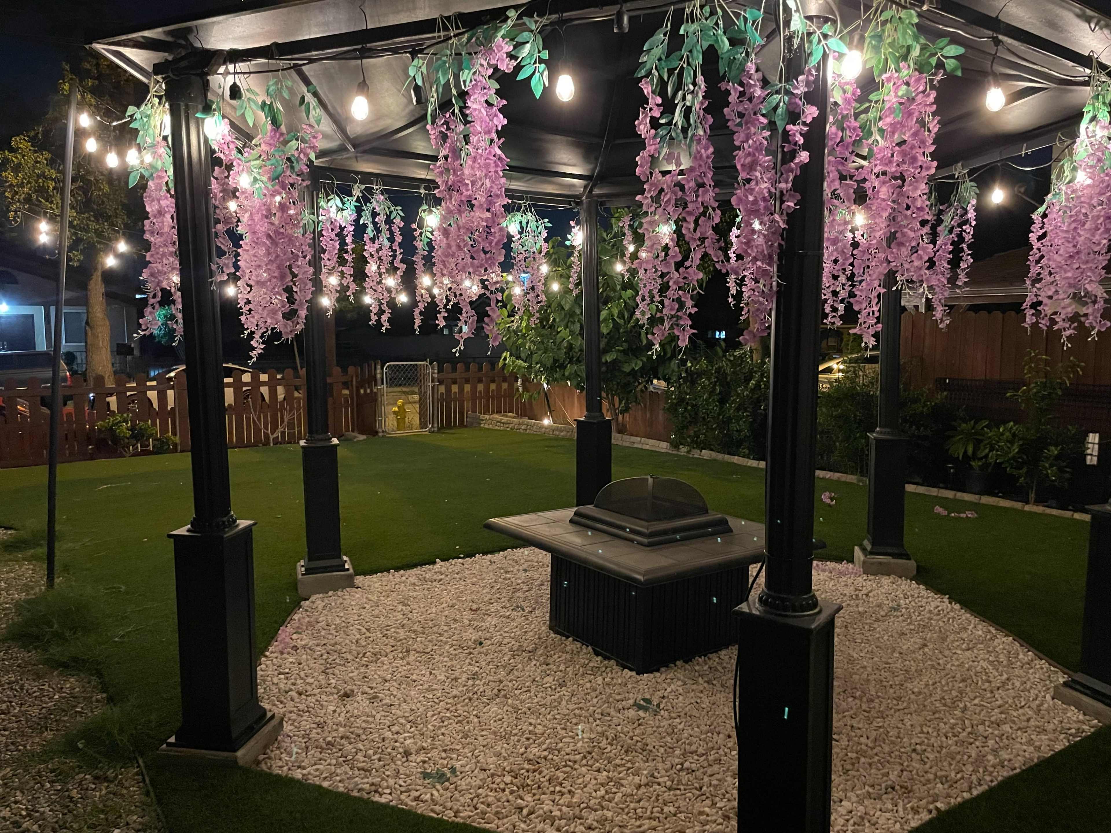 A gazebo adorned with purple flowers and string lights sits on a grassy area surrounded by decorative gravel.