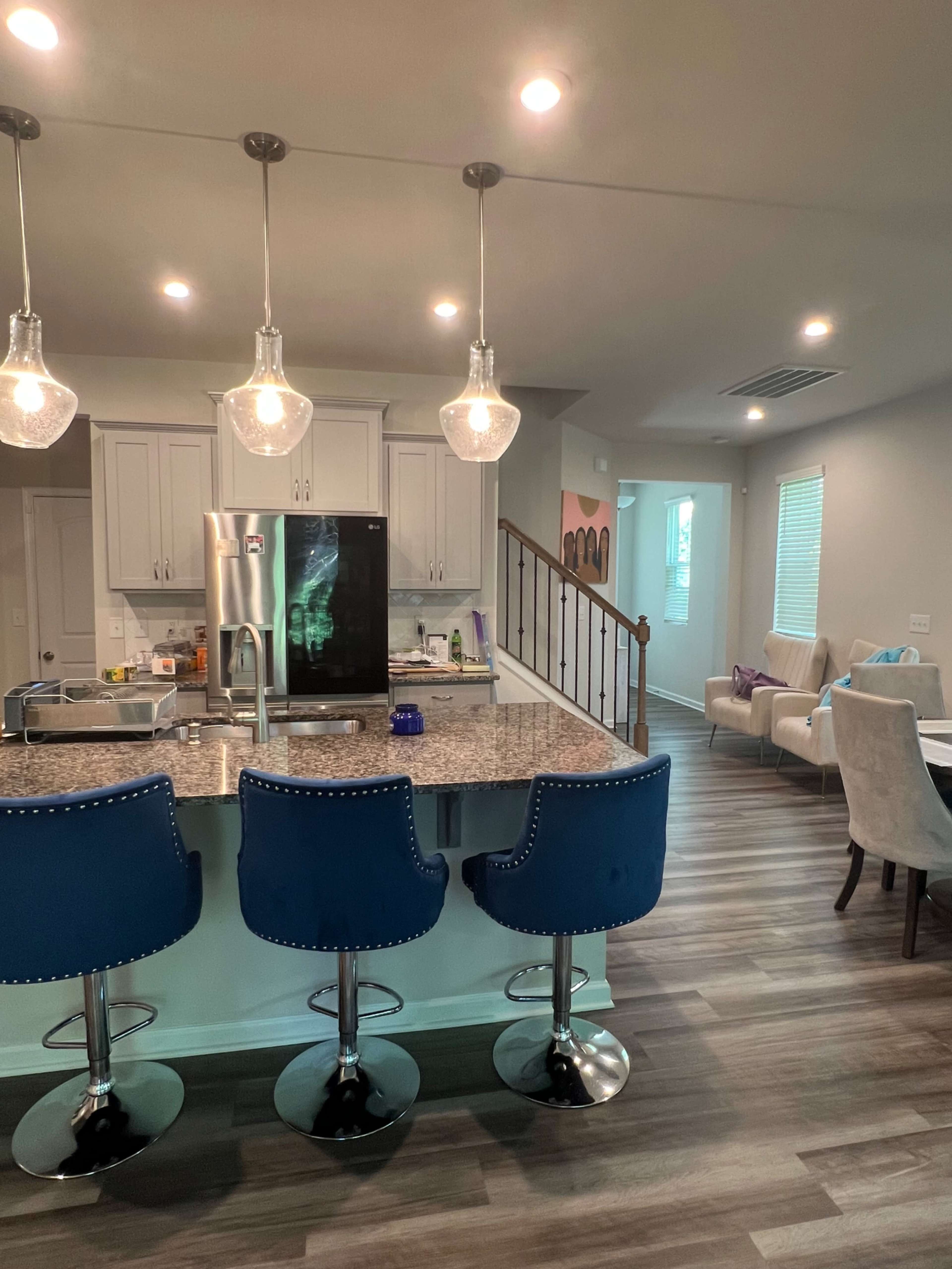 A modern kitchen features a granite island with three blue upholstered bar stools and a stainless steel refrigerator, while the adjacent living area includes a staircase and a cozy seating arrangement.