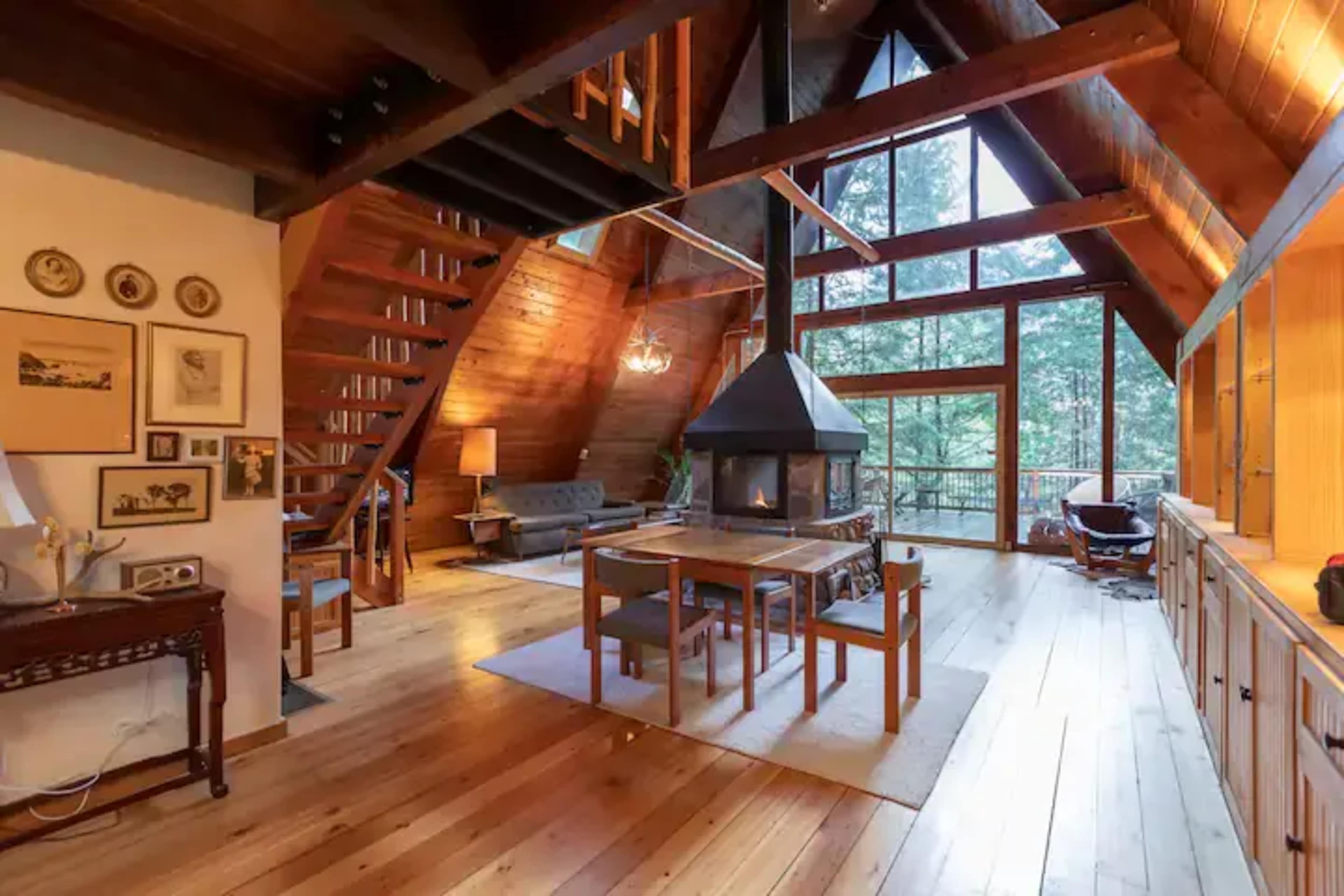 The interior of a wooden A-frame cabin features open spaces with a dining area, living space, and large windows overlooking trees.