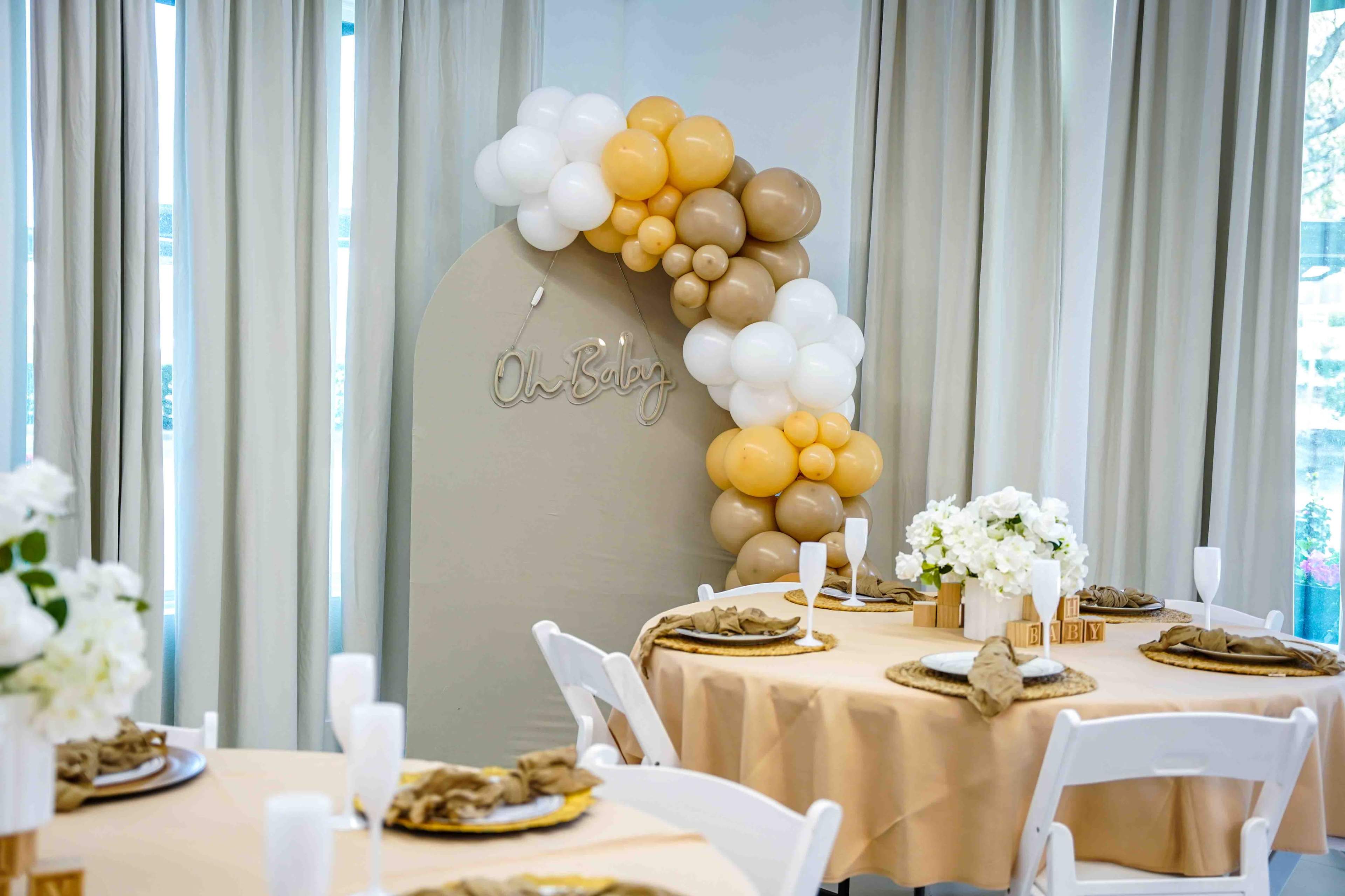 A decorated event space features tables set with neutral-colored tablecloths, floral centerpieces, and a balloon arch with a sign that reads "Oh Baby."