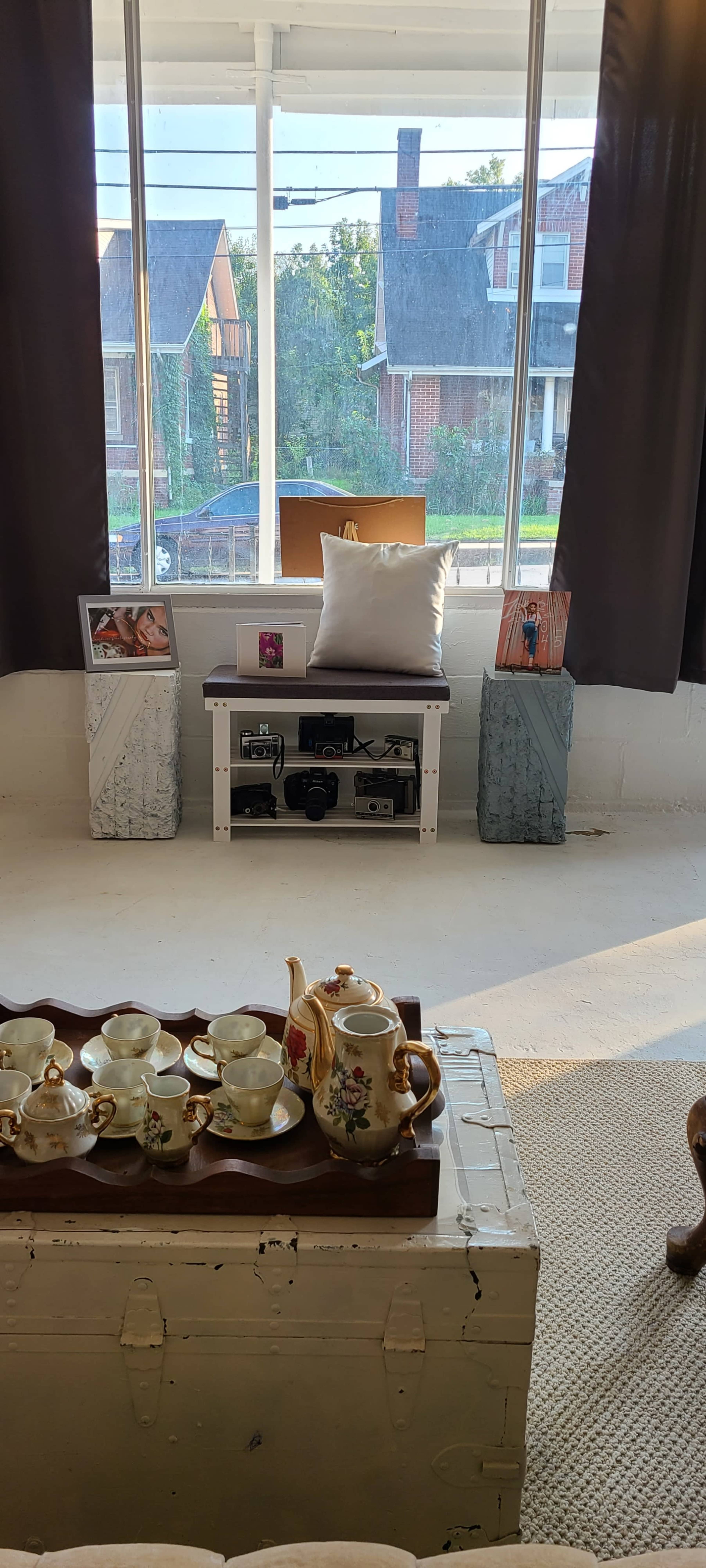 The scene shows a sunlit room with large windows, featuring a white table with a tea set, two decorative blocks on either side, and a photographic display on a low table.