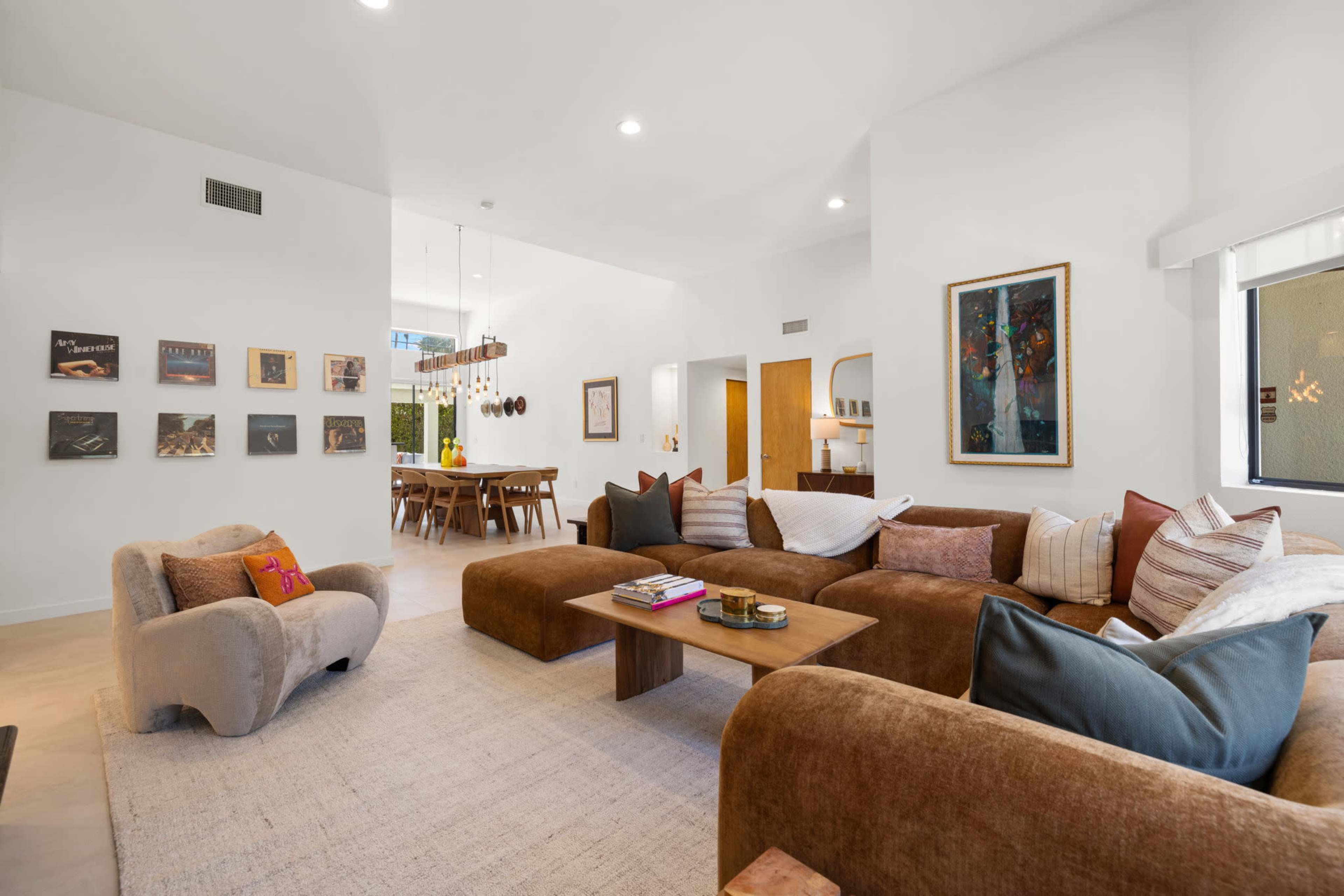 The scene shows a modern living room with a large sectional sofa, decorative pillows, and artwork on the walls, leading into a dining area with a wooden table and chairs.
