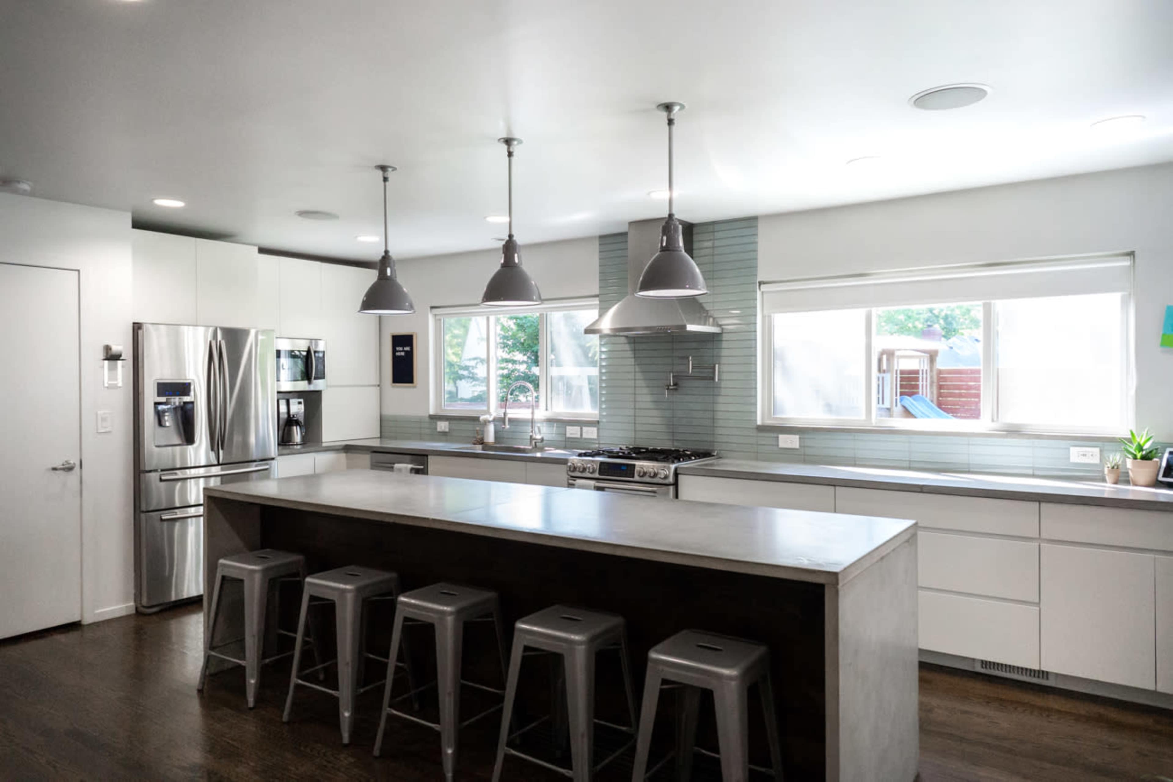 The modern kitchen features a central concrete island with metal stools, stainless steel appliances, and large windows that allow natural light to fill the space.