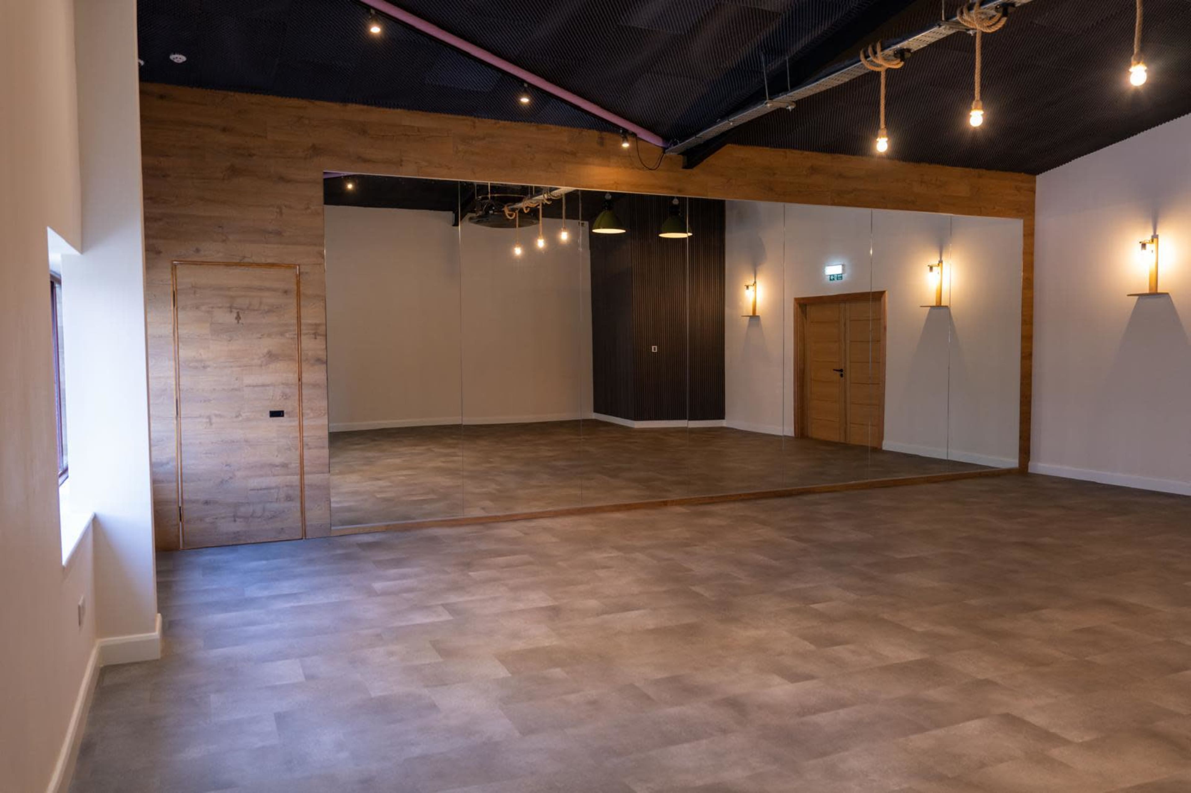 The image shows a spacious, well-lit interior room with wooden accents, a mirrored wall, and simple lighting fixtures.