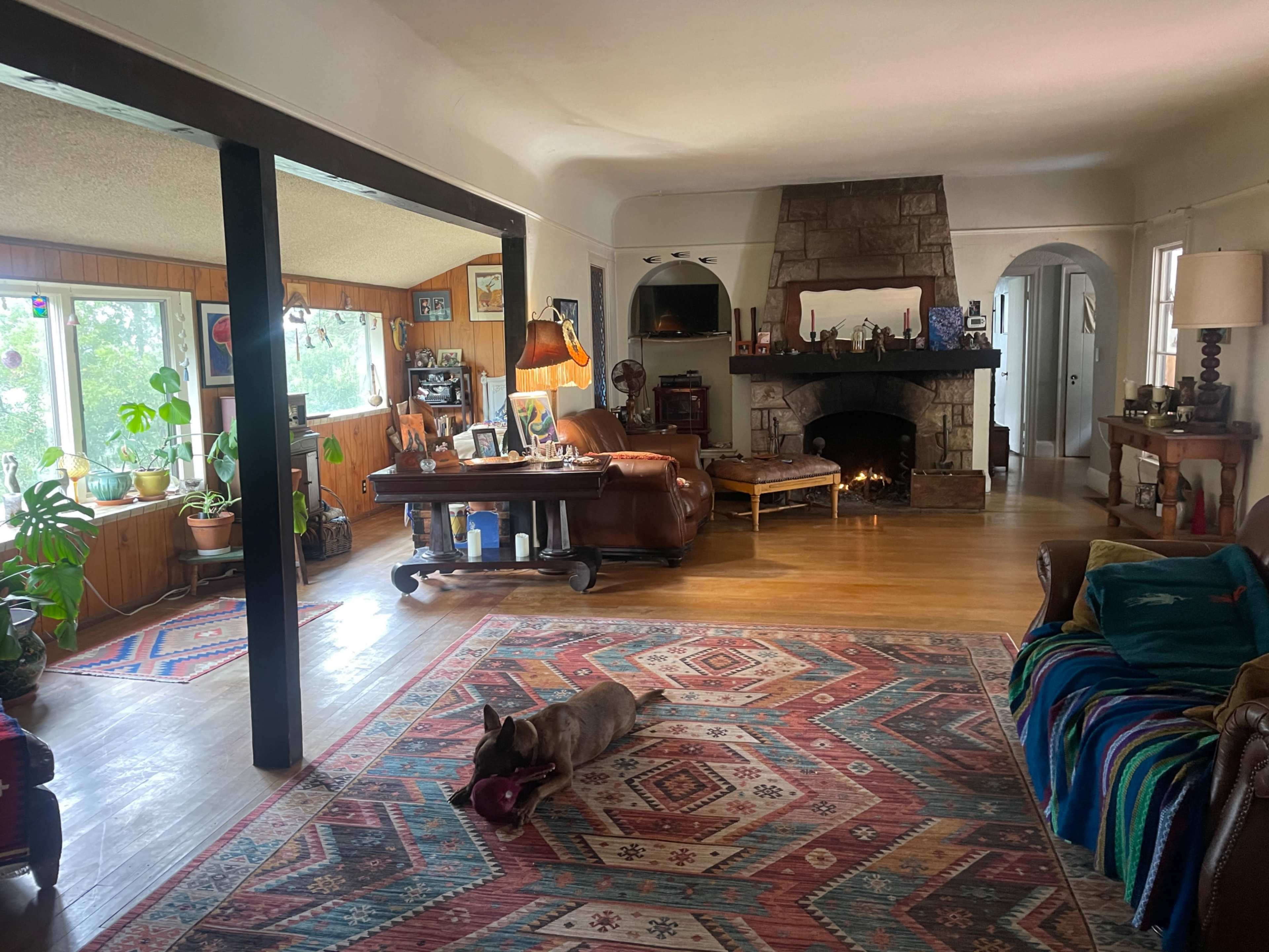 The image shows a cozy living room with a large stone fireplace, a rug on the hardwood floor, and a dog lying on the rug near a wooden coffee table.