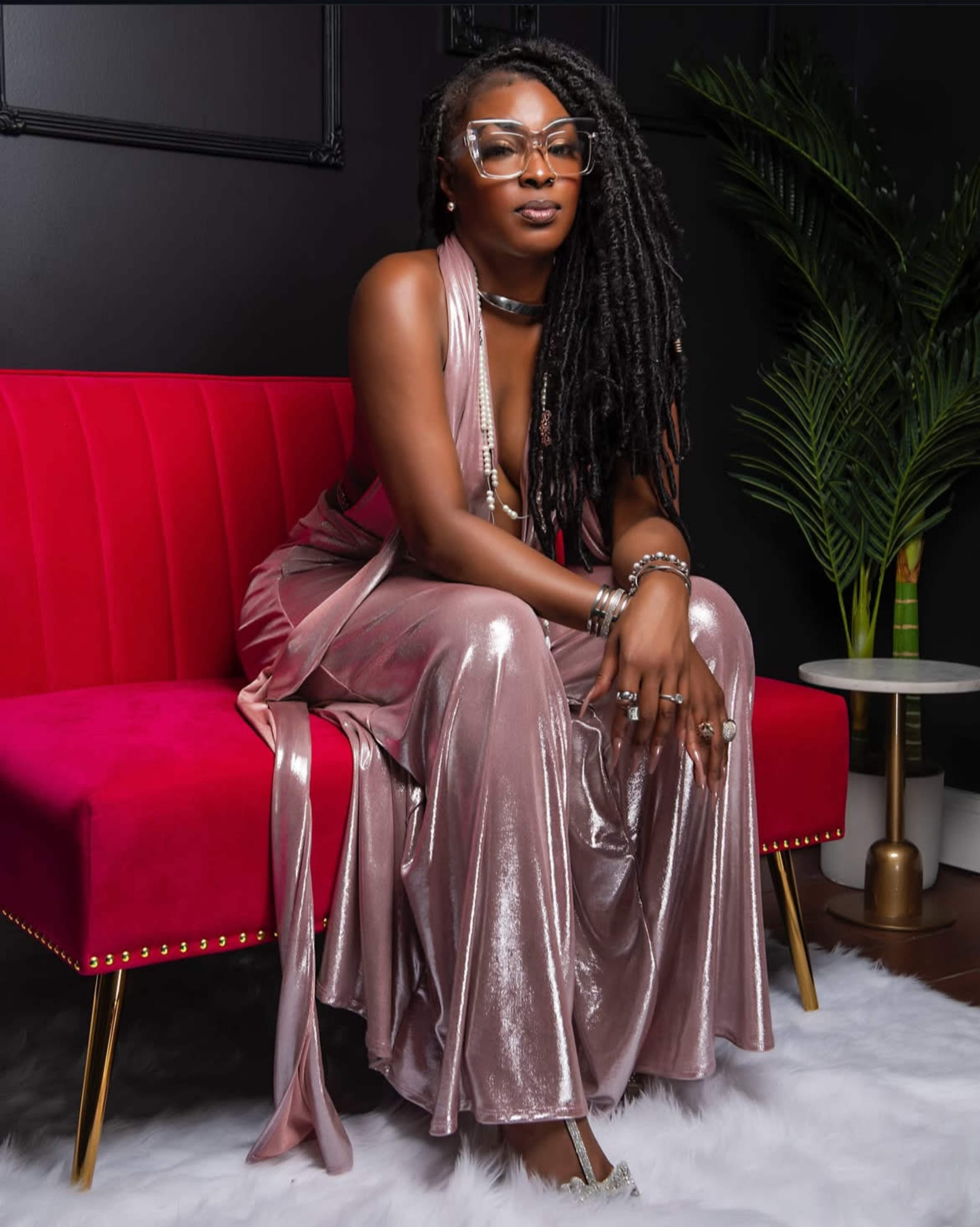 A woman with long, curly hair is seated on a pink velvet couch, wearing a shimmering, floor-length dress and accessorized with glasses and jewelry.