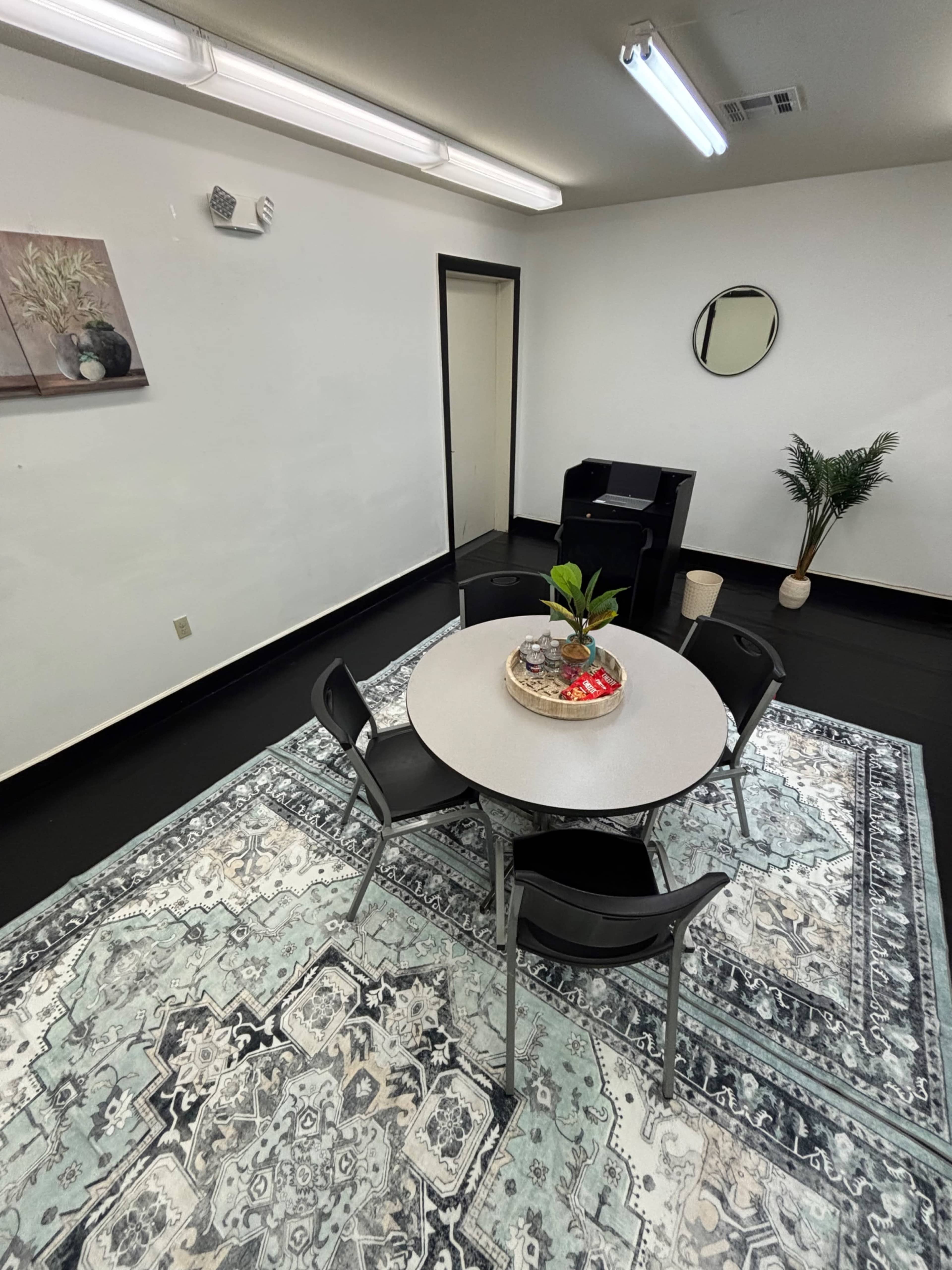 The image shows a simple meeting room with a round table surrounded by four chairs on a patterned rug, and a black armchair in one corner.