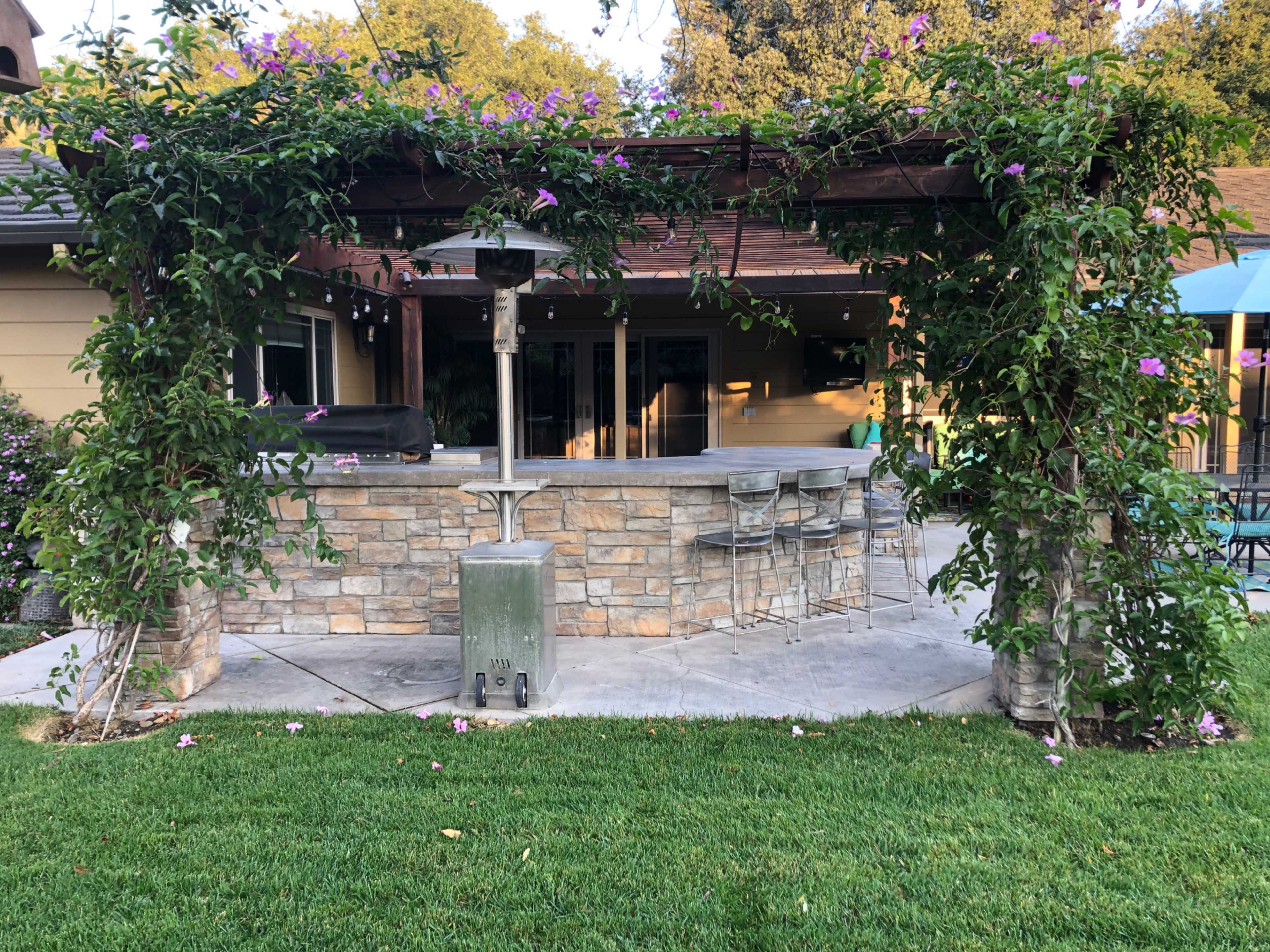 A stone outdoor bar area with a pergola adorned with greenery and a few bar stools, set in a grassy yard.