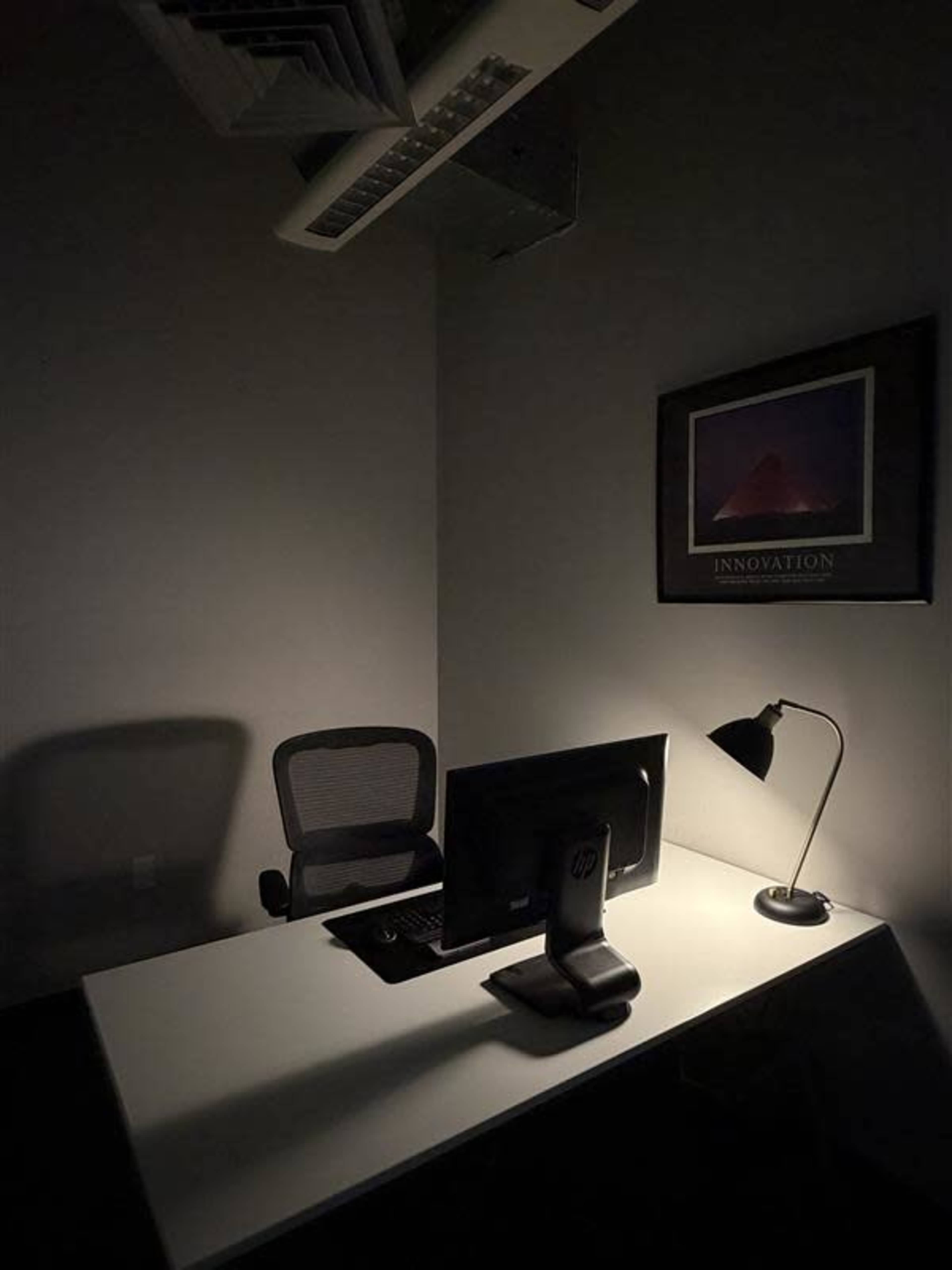 A dimly lit office features a desk with a computer monitor, a lamp, and a chair against a gray wall.