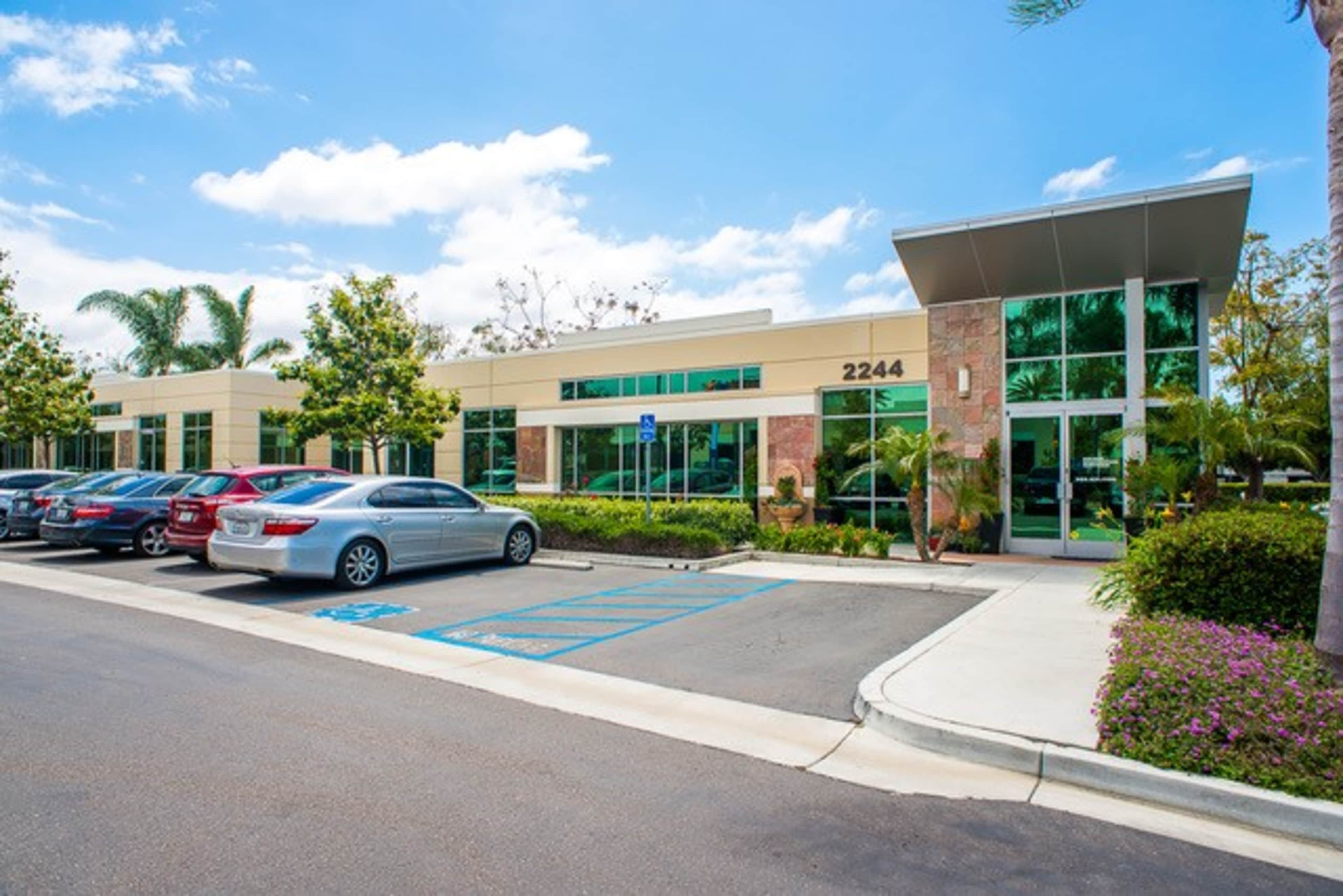 The image shows a modern office building with large glass windows and a parking lot filled with vehicles.
