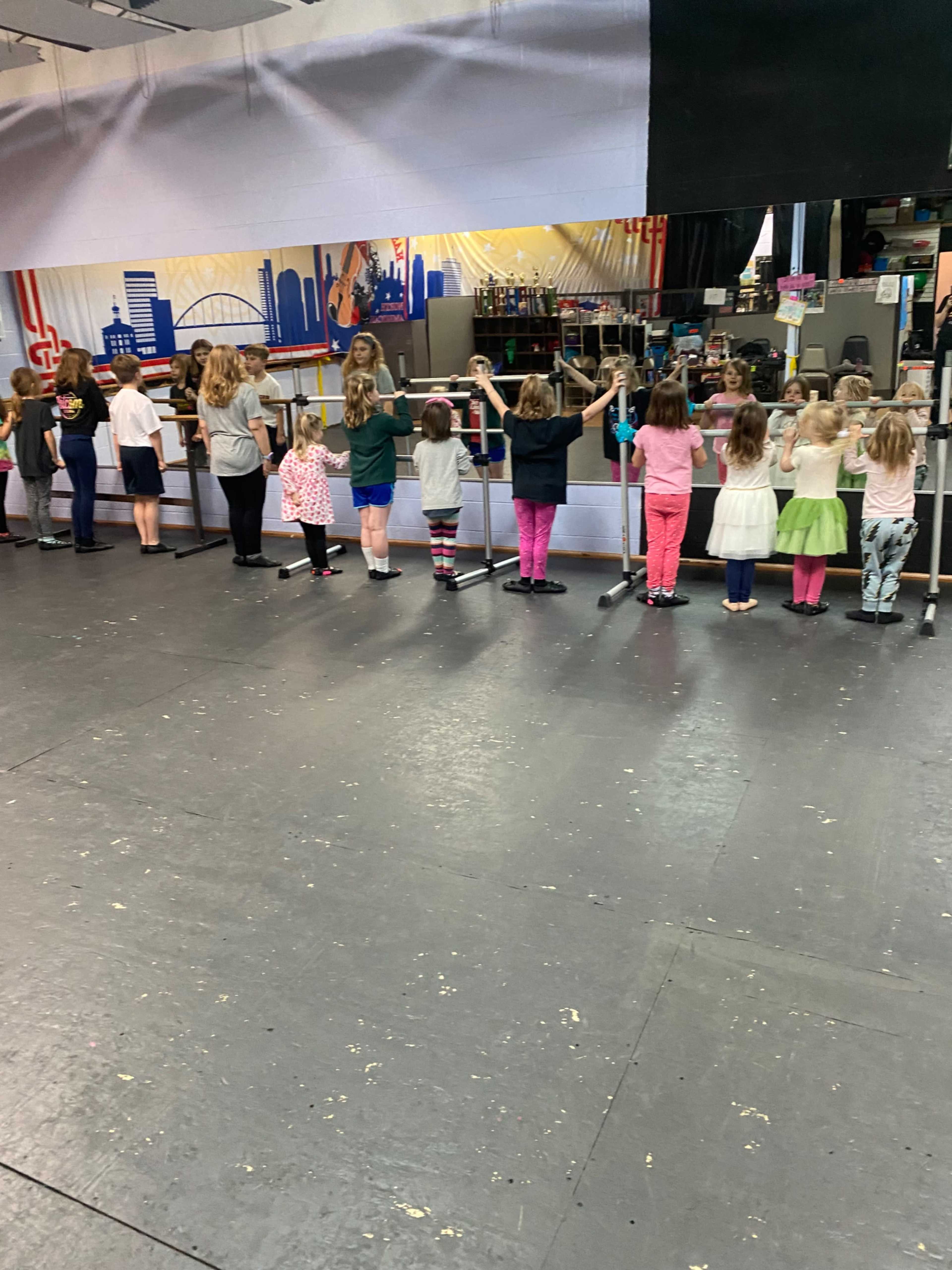 A group of children stands in line at ballet practice, holding onto a ballet barre in a dance studio.