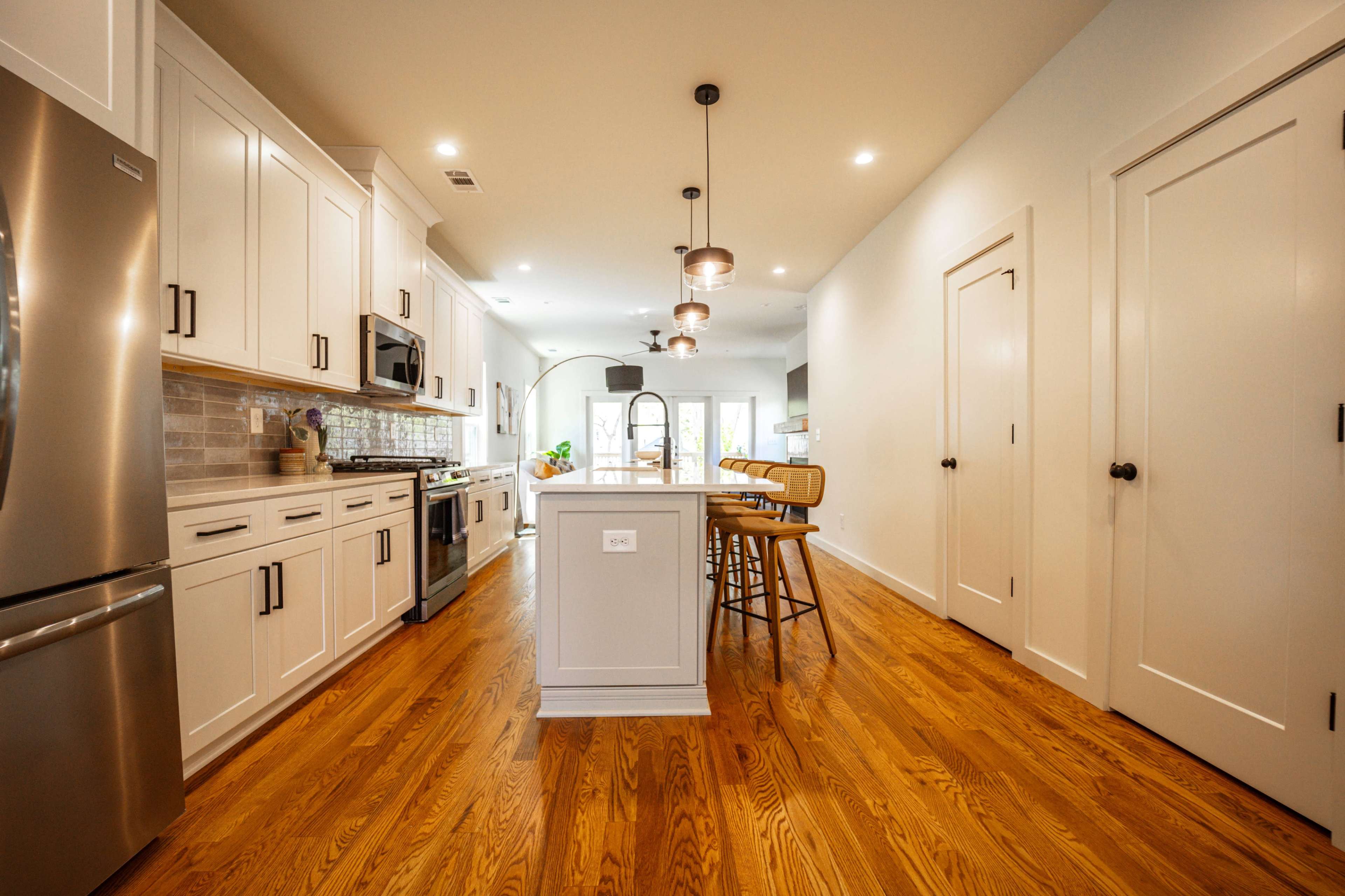 The image depicts a modern kitchen featuring white cabinetry, stainless steel appliances, a central island with bar stools, and wooden flooring.