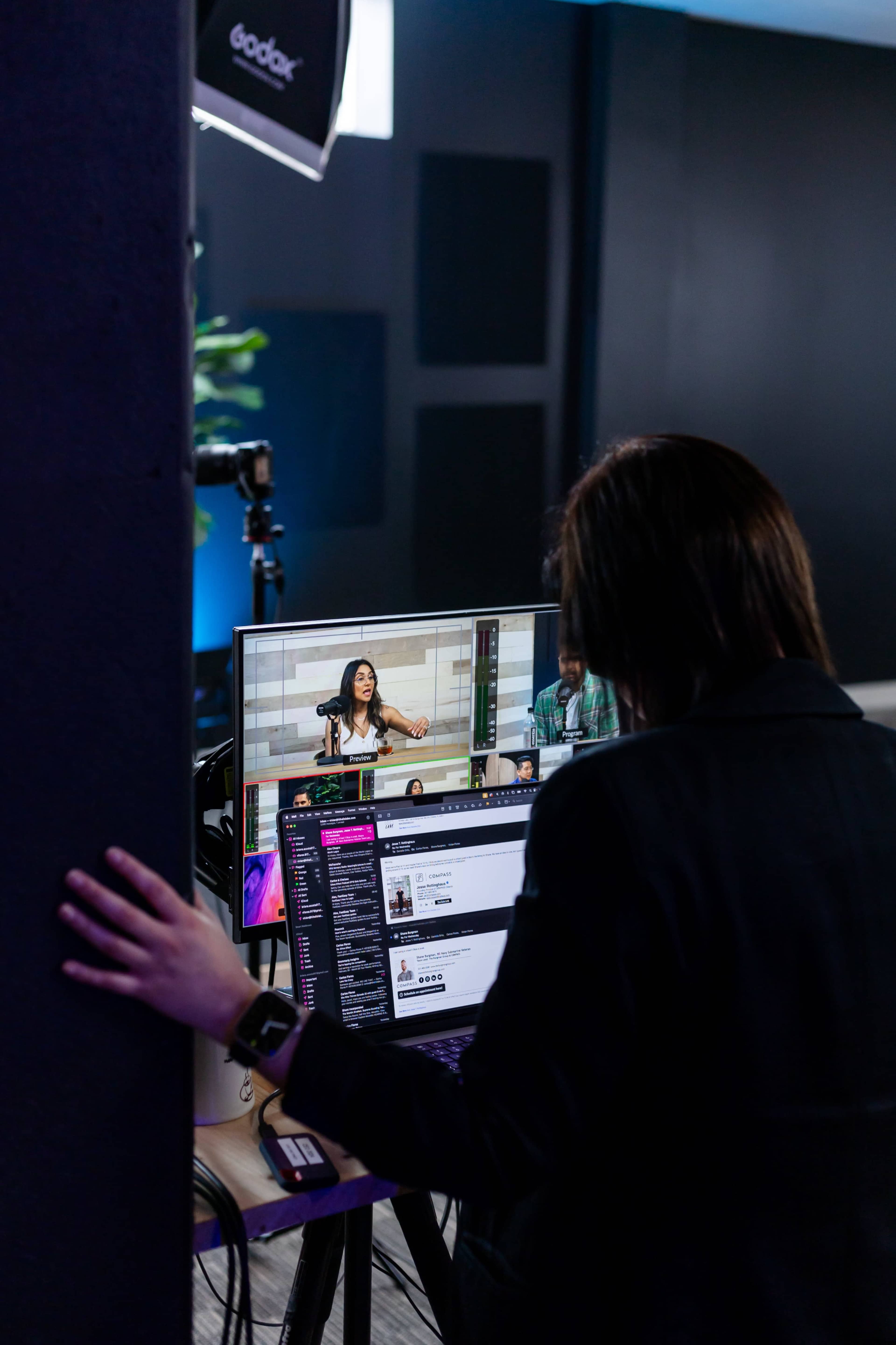 A person is seated at a desk with a laptop, watching a video feed of a woman speaking on a large screen in a studio setting.