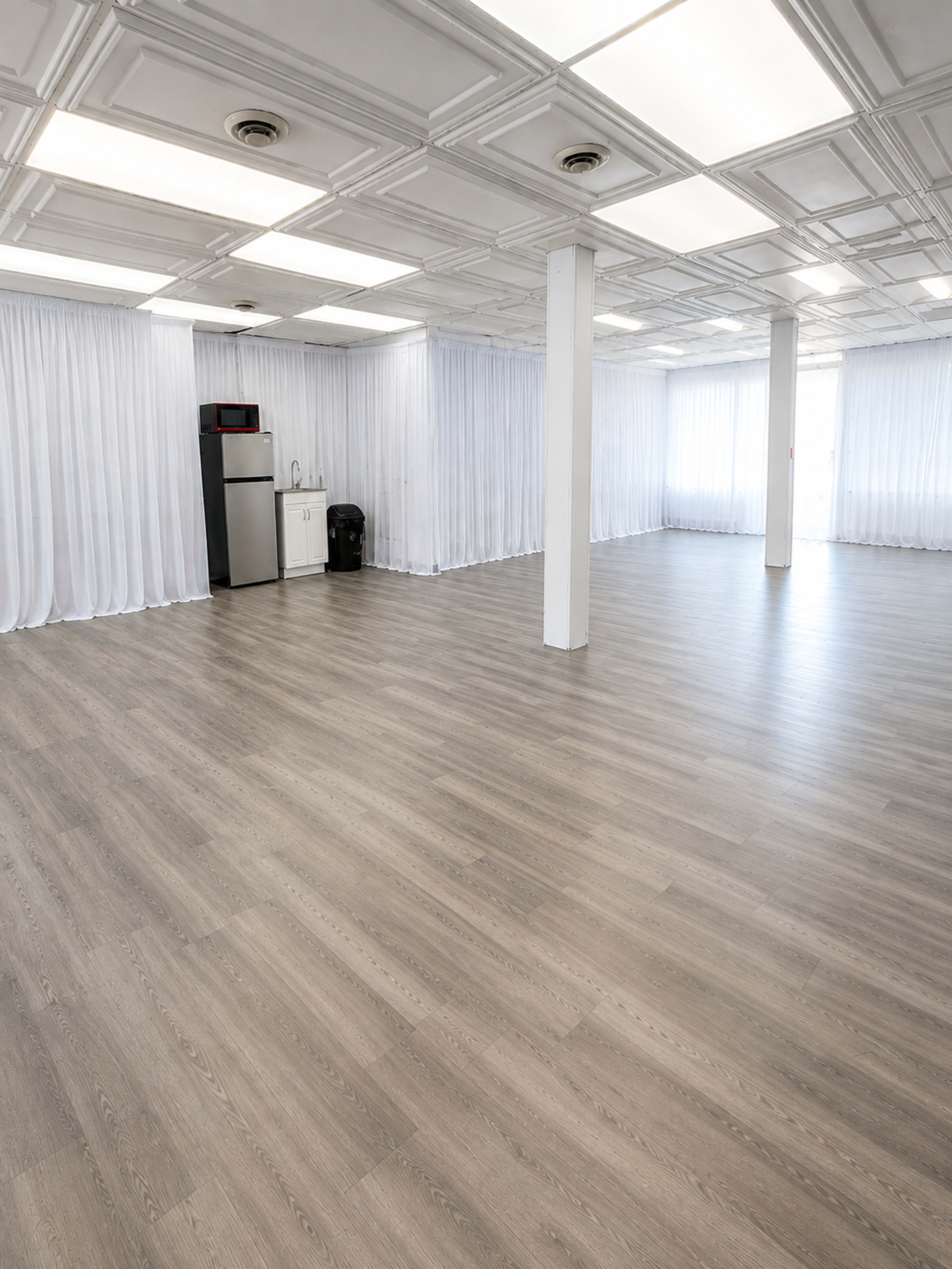 The image shows an empty room with light wooden flooring, white curtains, and a kitchenette area including a refrigerator and a trash can.