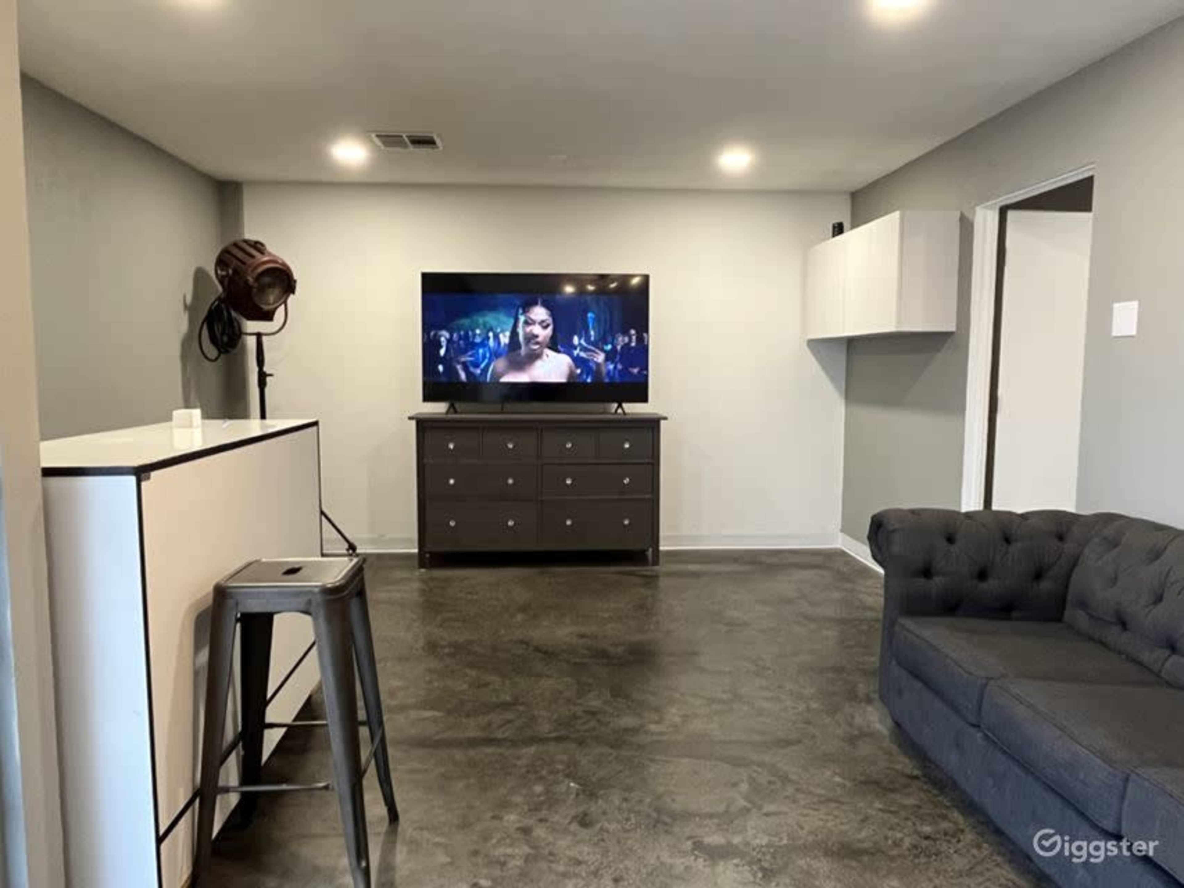 A modern living room with a large television displaying a scene from a movie, a dark gray couch, a dresser, and a barstool near a white cabinet.