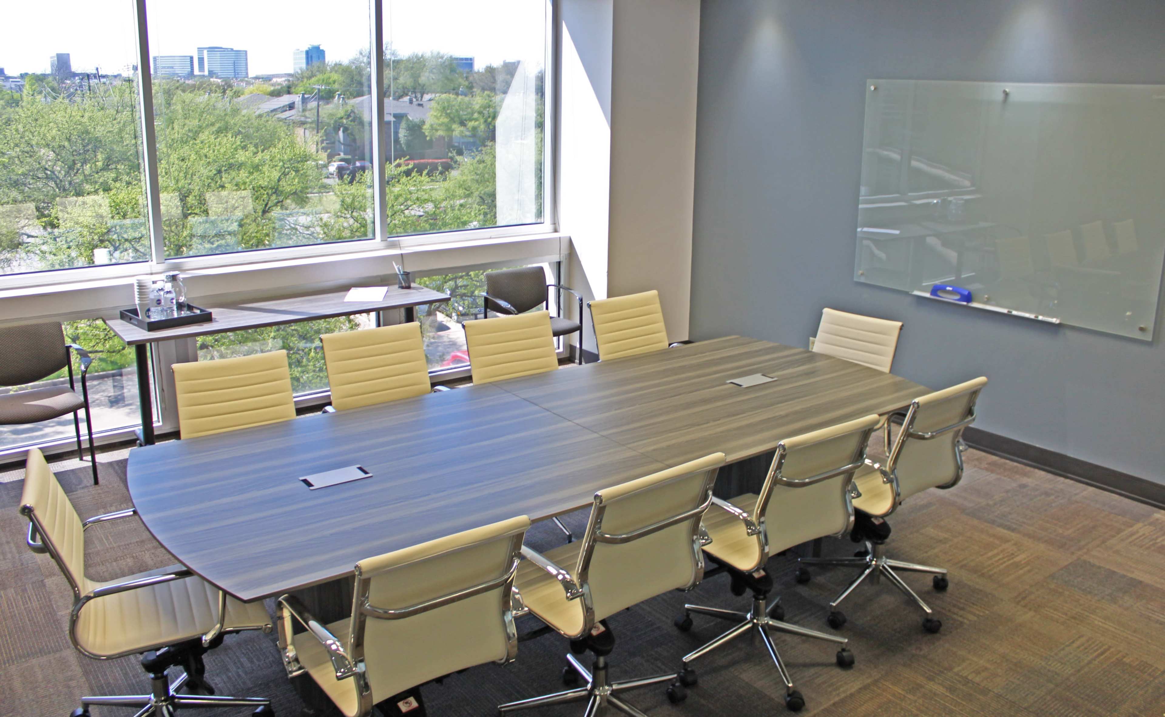 A large conference room features a long table surrounded by several chairs, with windows offering a view of greenery and buildings outside.