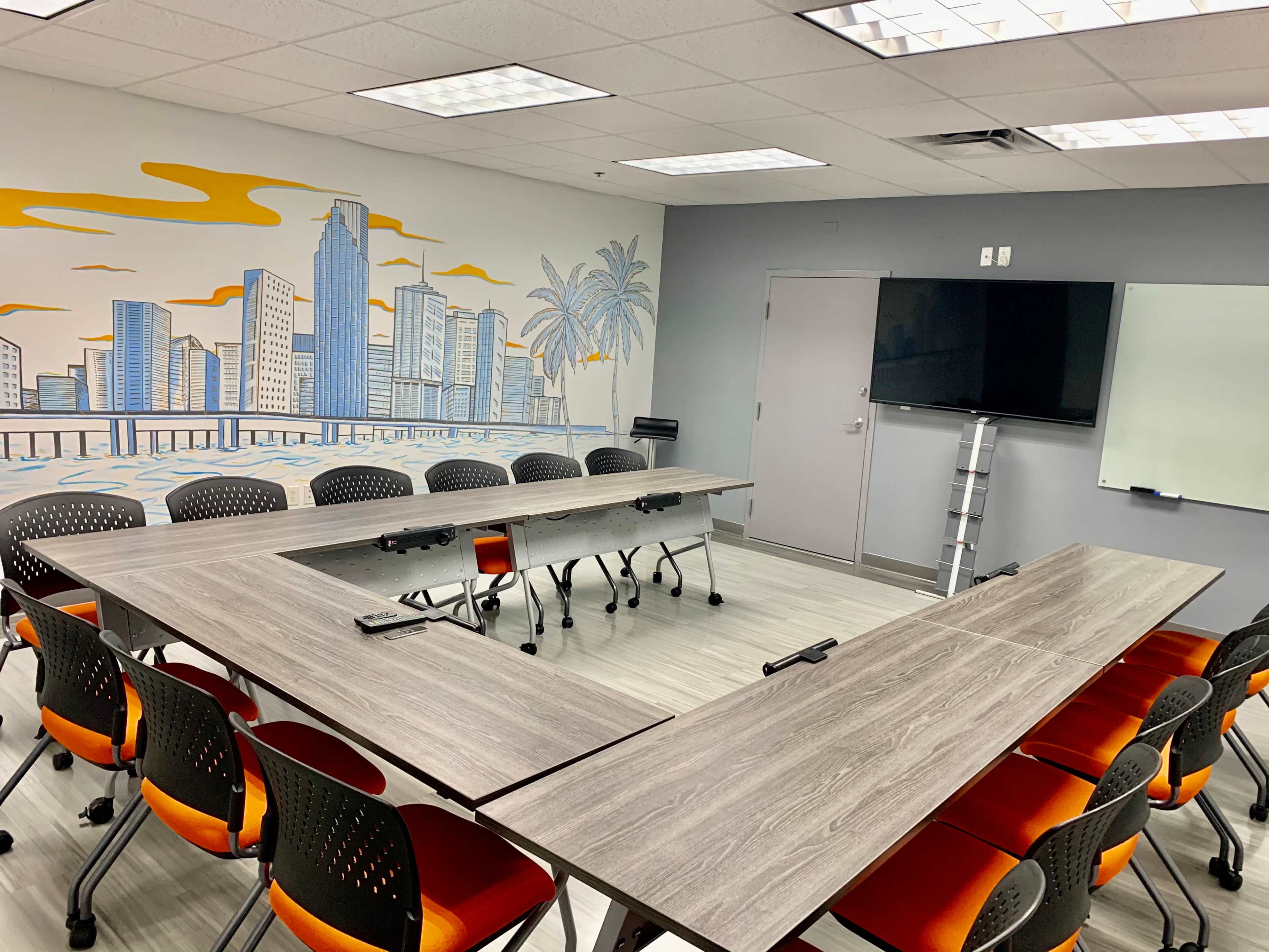 A conference room features a U-shaped arrangement of tables with orange and black chairs and a wall mural of a city skyline.
