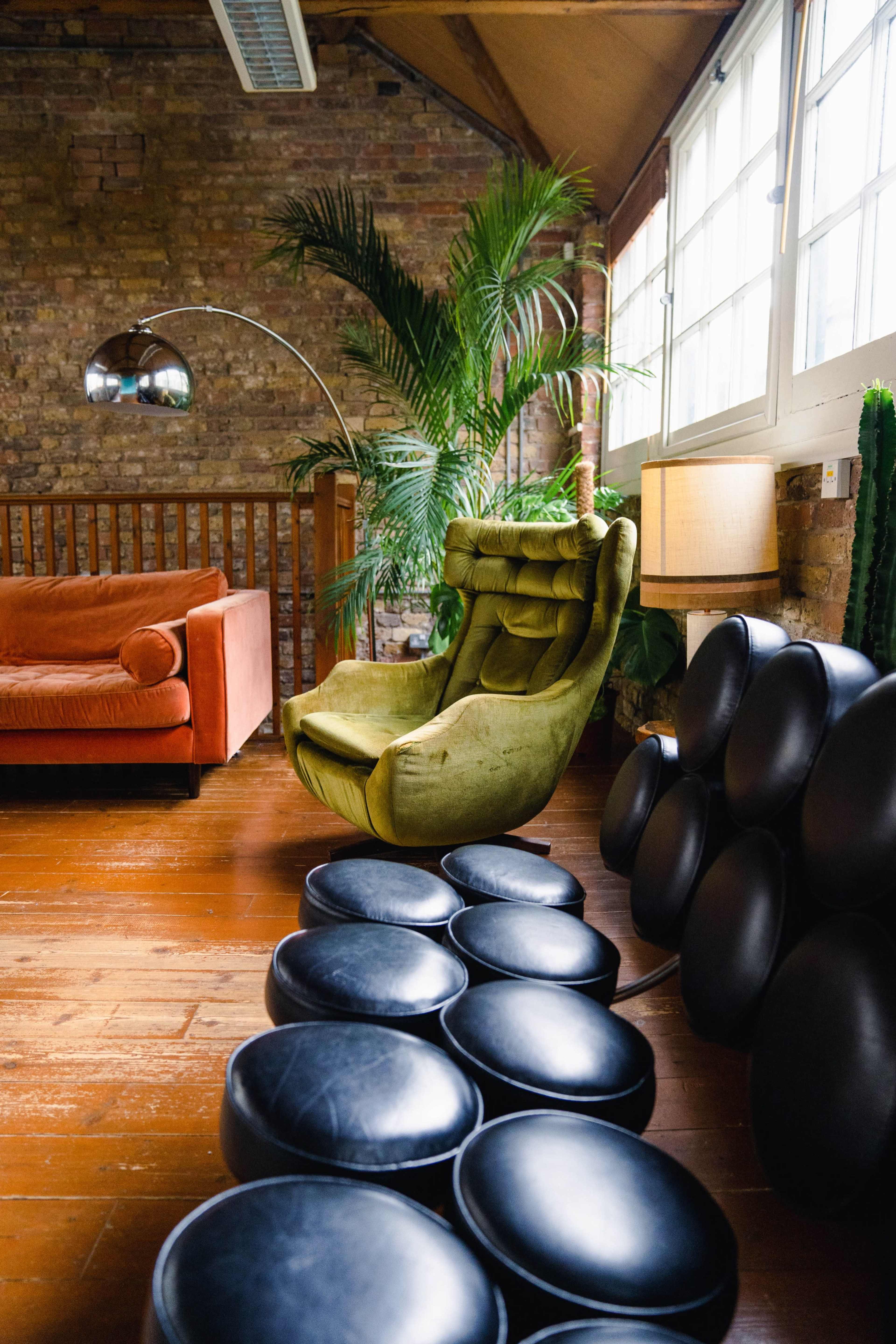 The room features a green lounge chair, a red sofa, and several round black ottomans, along with plants and large windows.