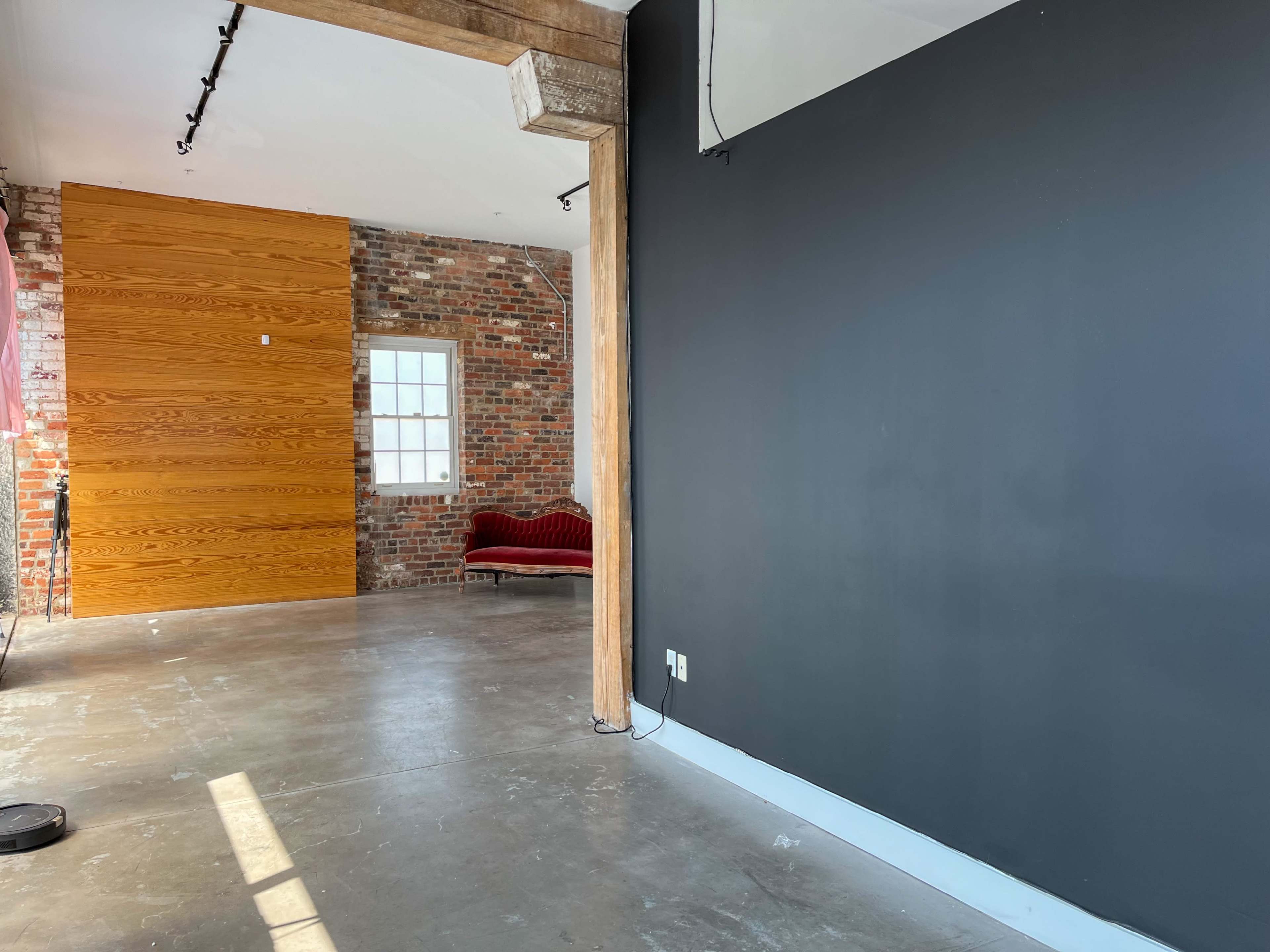 The image shows an interior space with a dark wall, a section of exposed brick, a wooden wall panel, large windows, and a red couch in the background.