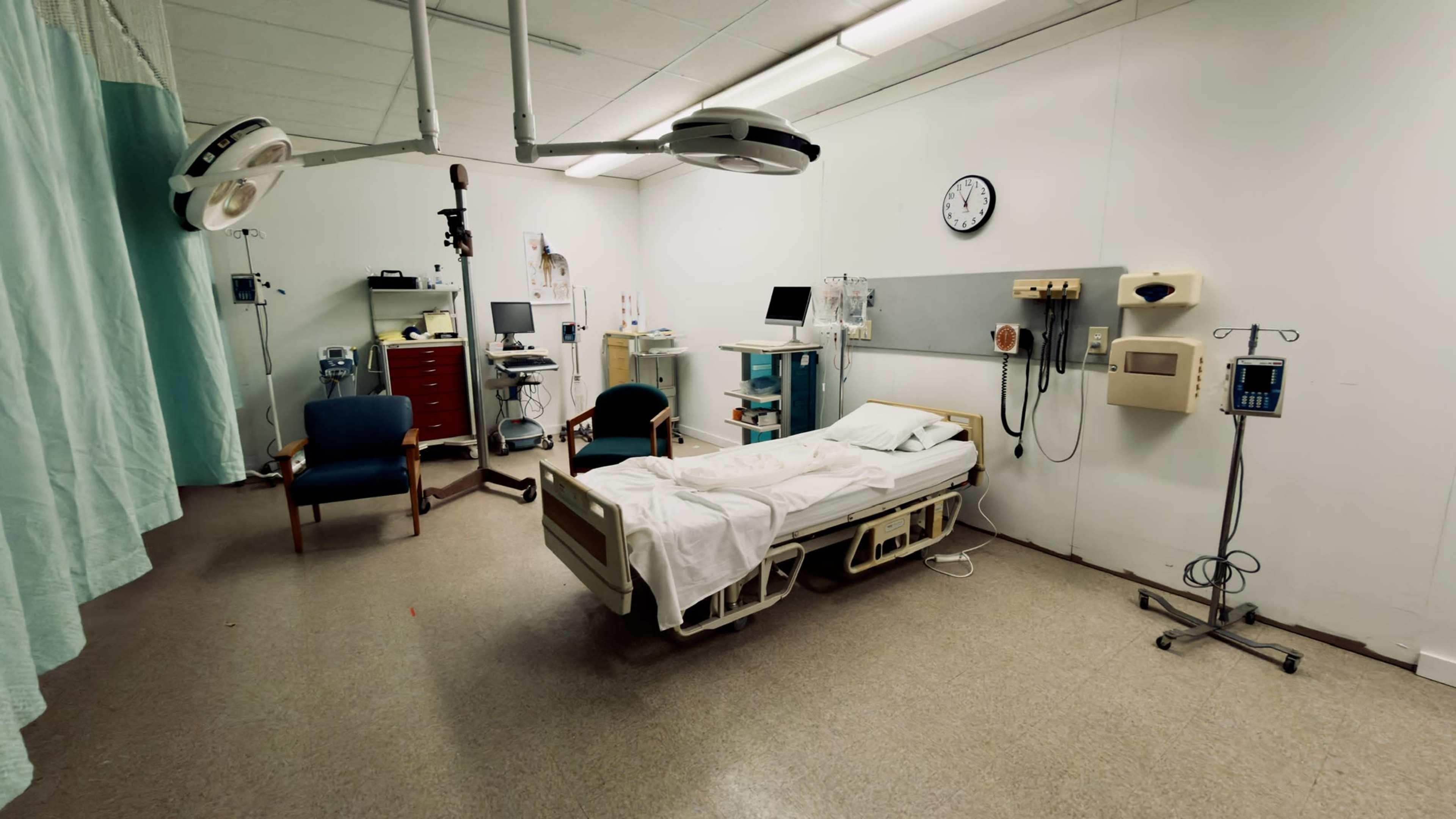 The image shows a hospital room with a bed, medical equipment, a clock, and chairs arranged along the walls.