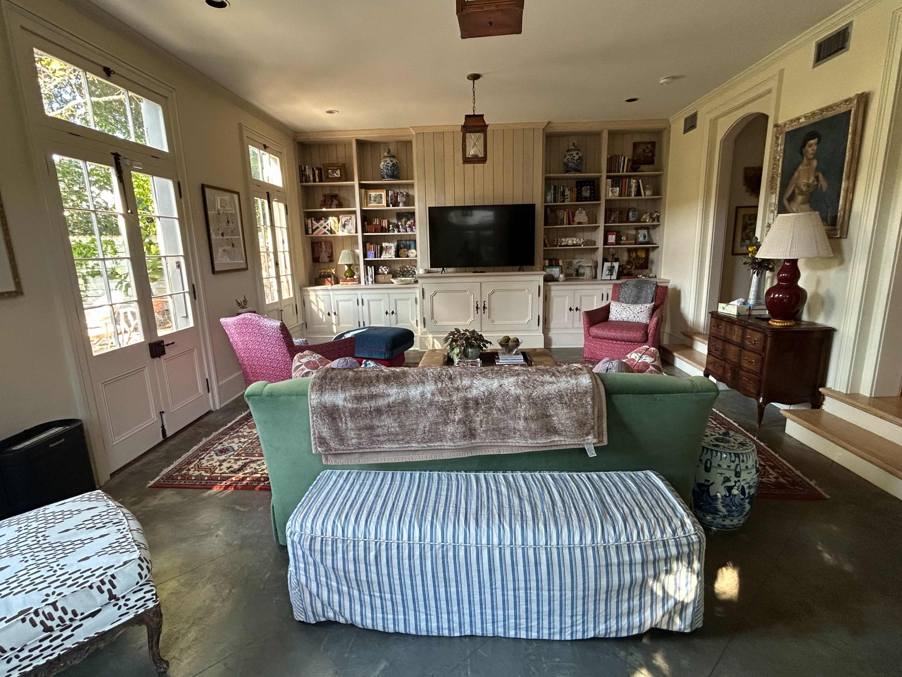 A cozy living room features a sofa, a striped ottoman, and armchairs arranged around a television on a built-in cabinet, with bookshelves and windows providing ample light.