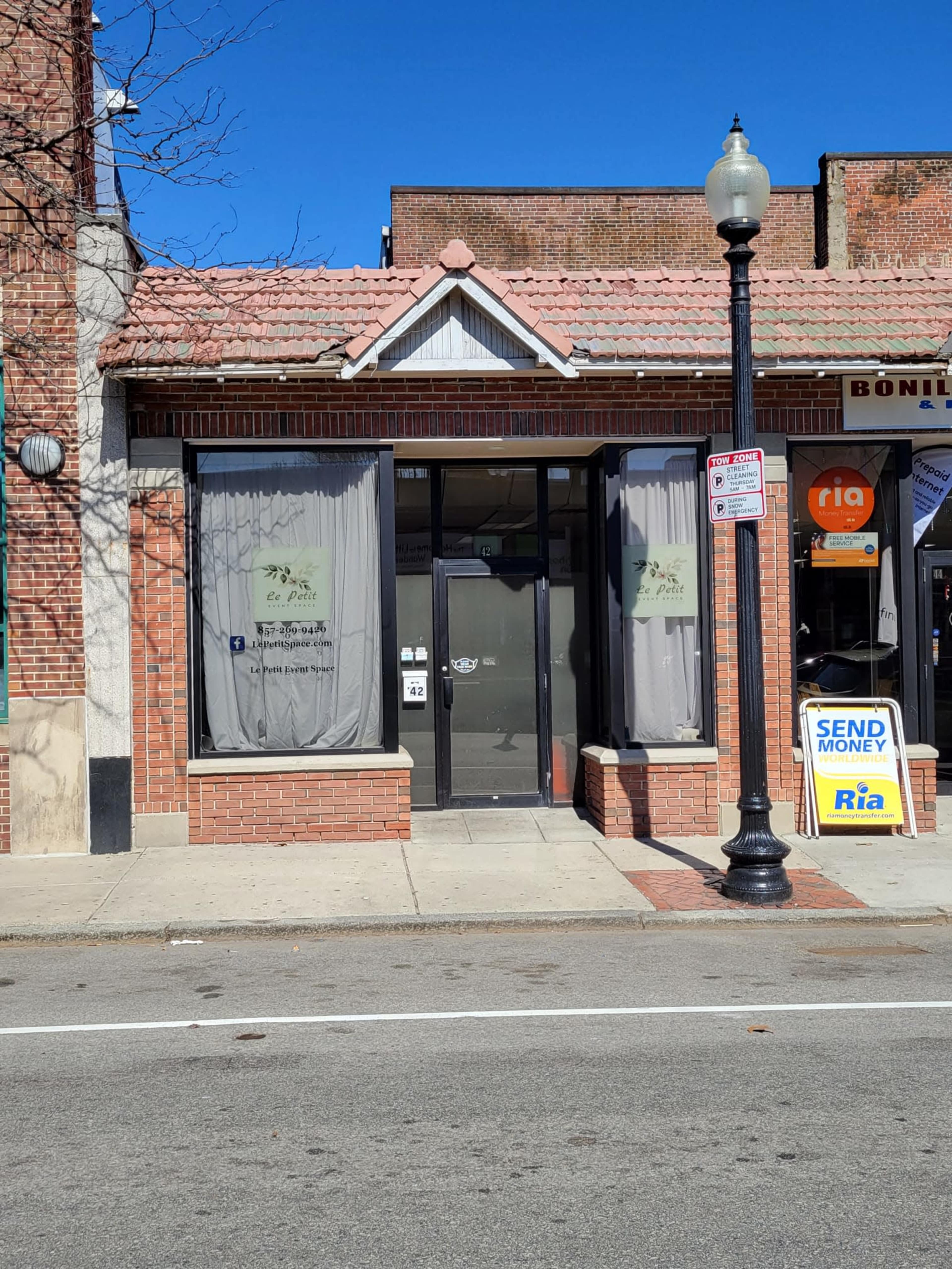 The image shows the entrance of a storefront with large windows covered by sheer curtains, located on a brick building in a commercial area.