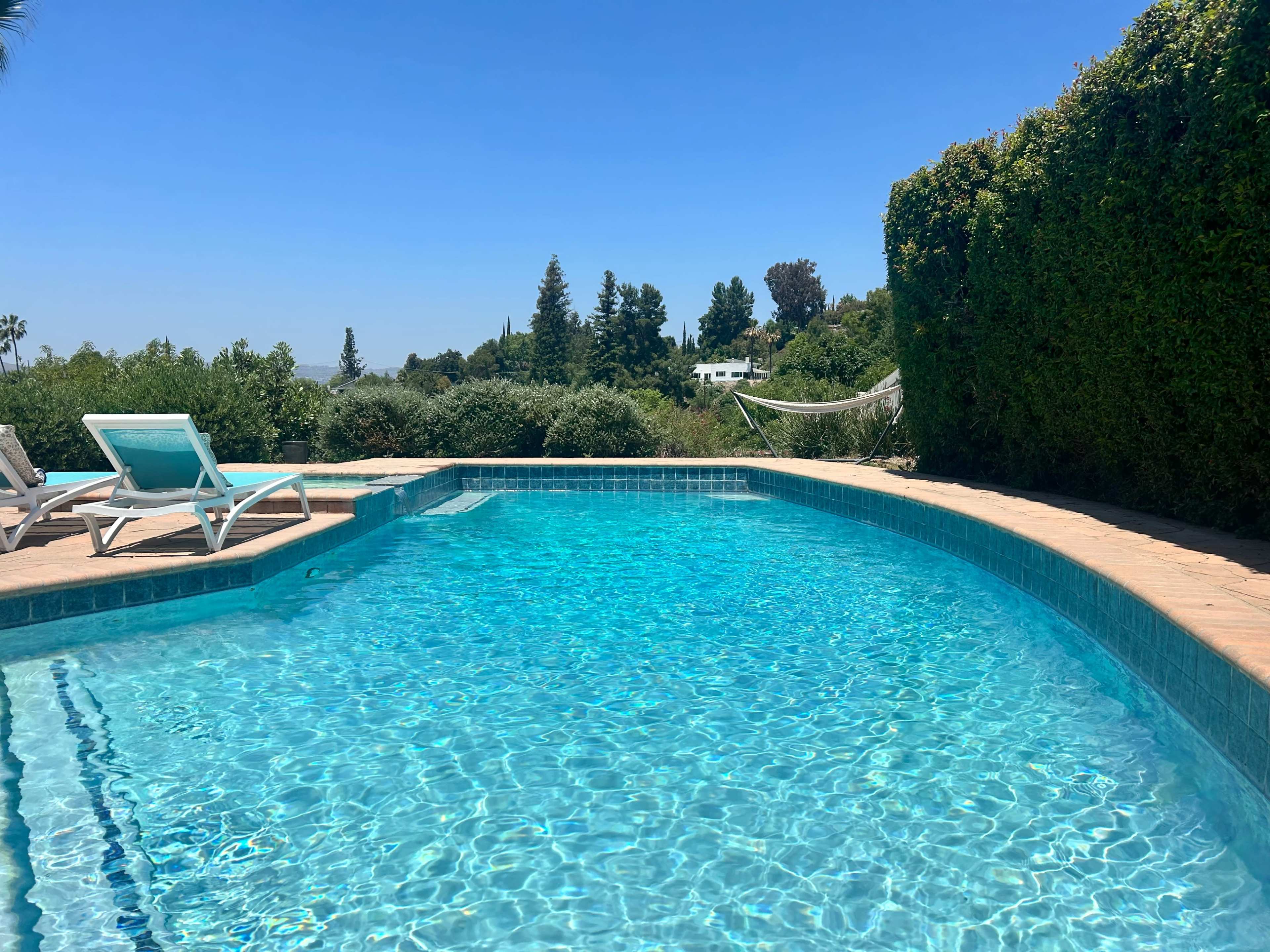 A clear blue swimming pool is surrounded by lush greenery and lounge chairs under a bright sky.