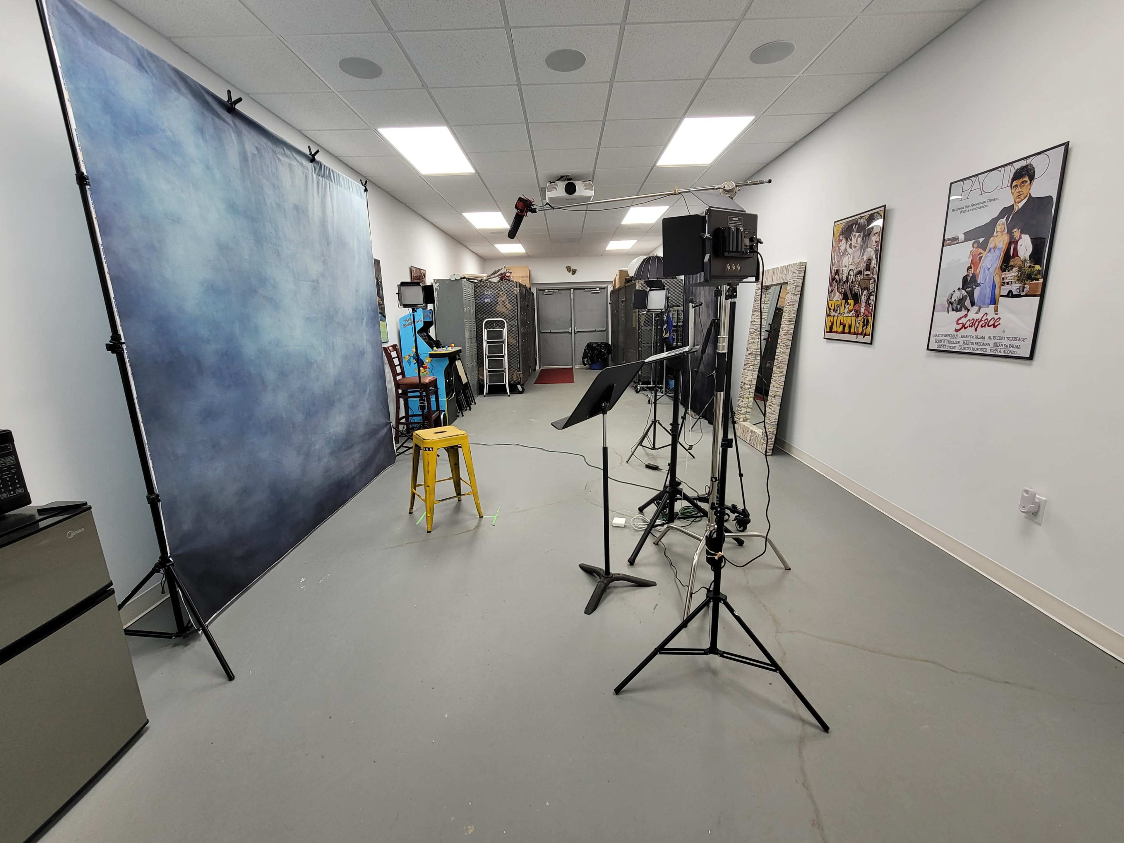 The image shows a well-lit studio space with a blue backdrop, several lighting equipment stands, a microphone stand, and film posters on the walls.
