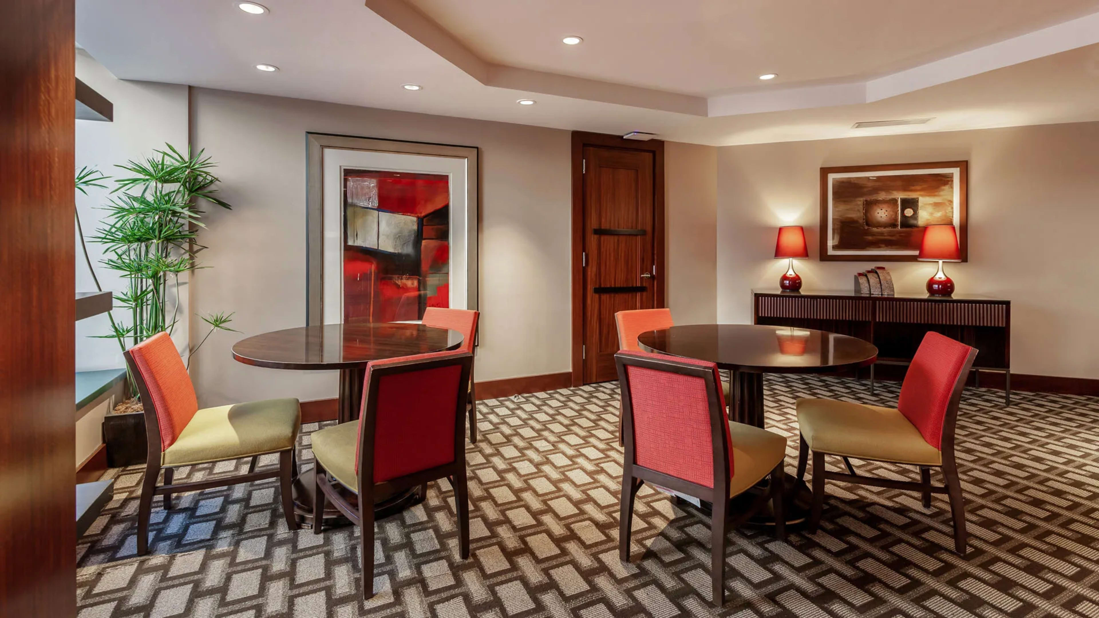 The image shows a modern lounge area featuring two round tables surrounded by chairs, a patterned carpet, and decorative artwork on the walls.