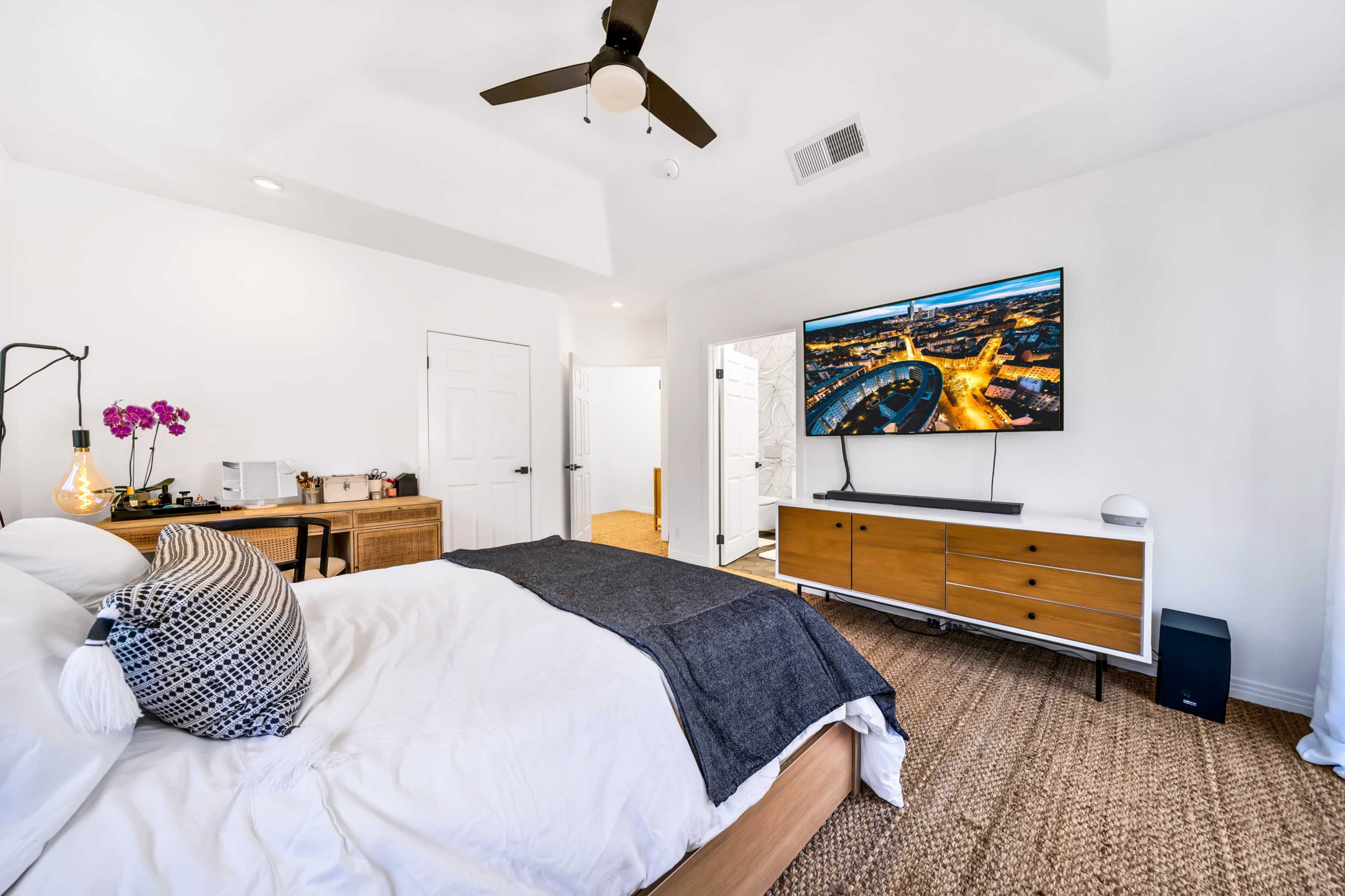 A modern bedroom features a large bed with white bedding, a flat-screen TV mounted on the wall, and a wooden dresser with a TV stand.