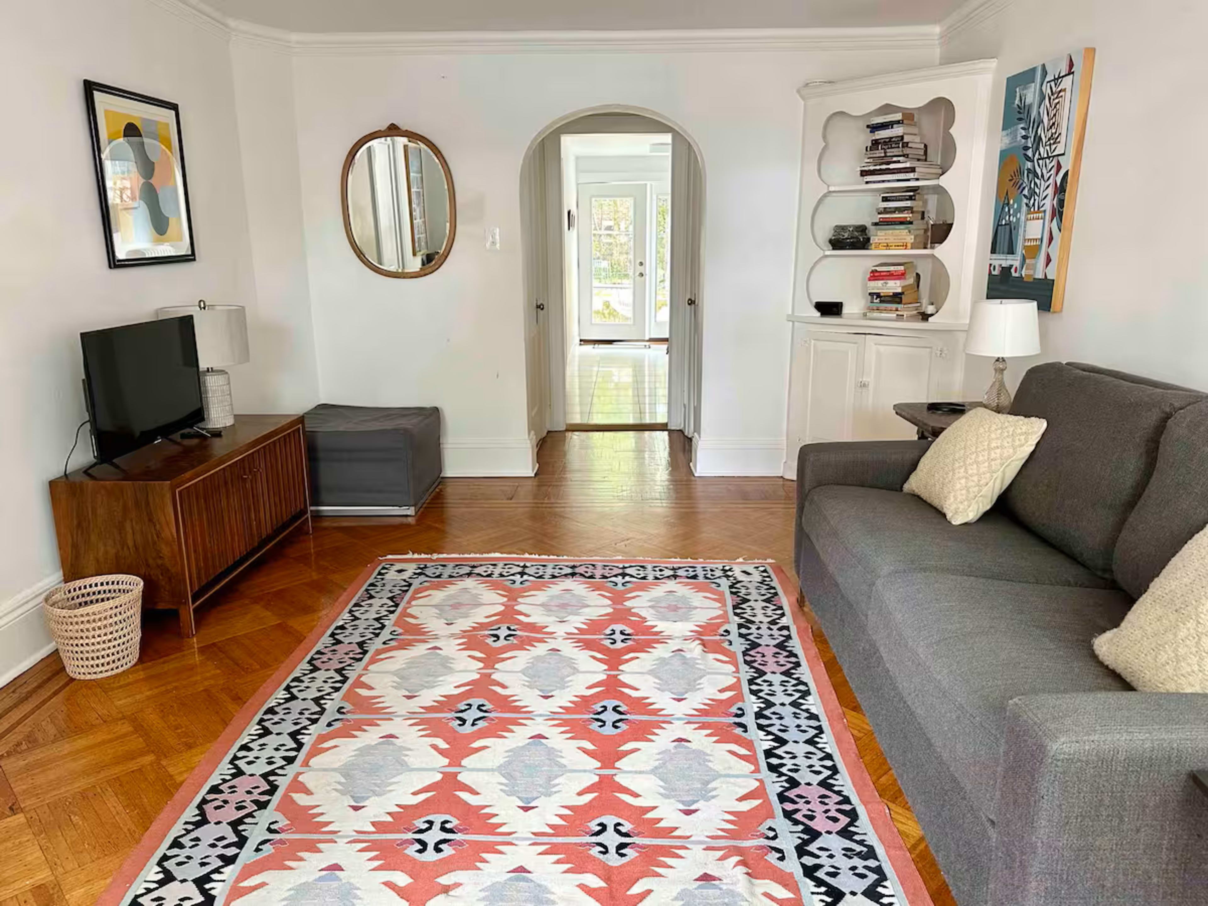 The living room features a gray sofa, a patterned area rug, a television on a wooden stand, and a doorway leading to a bright hallway.