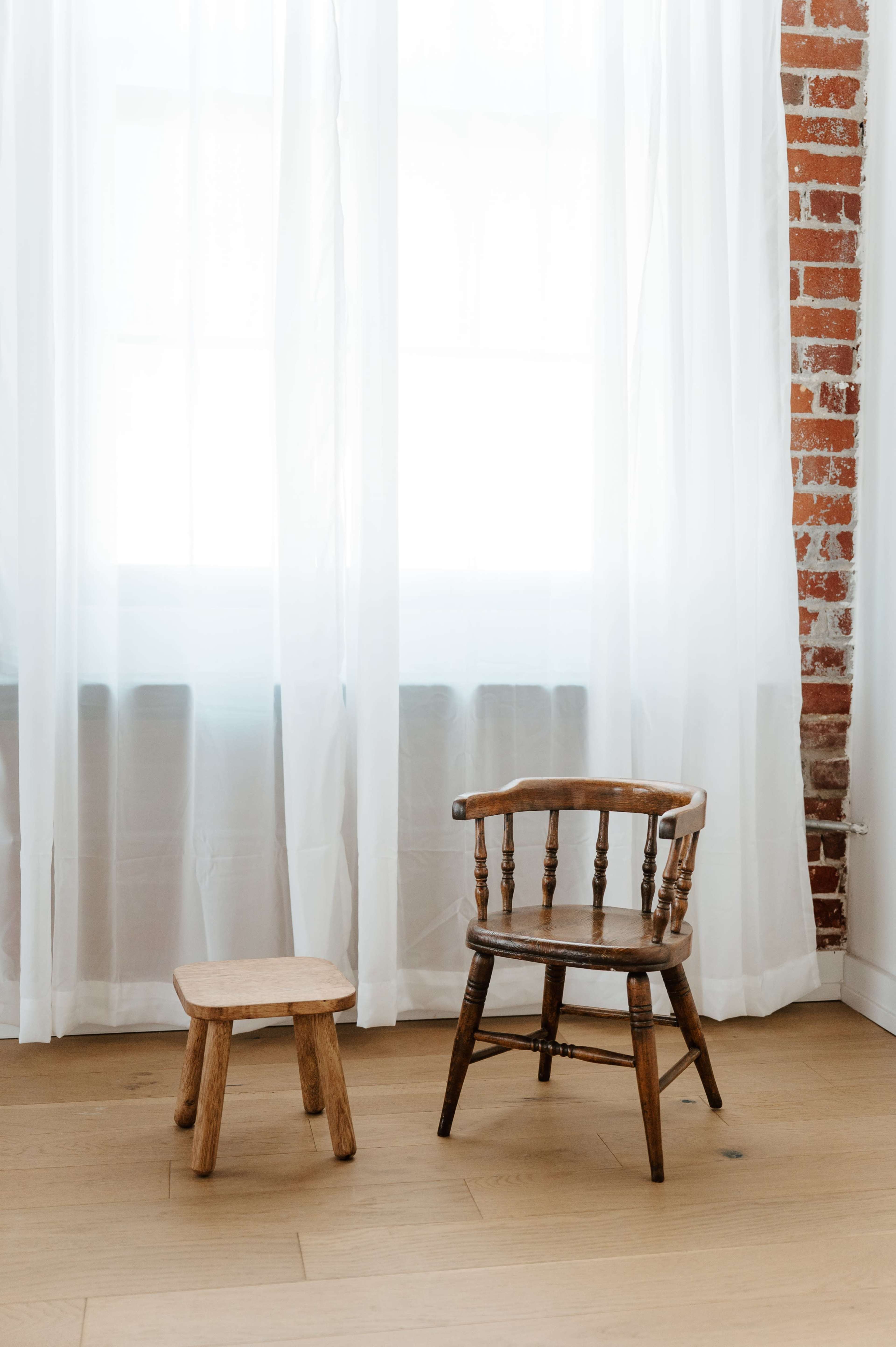 The image features a wooden chair and a small stool placed next to each other beneath a window with sheer white curtains.