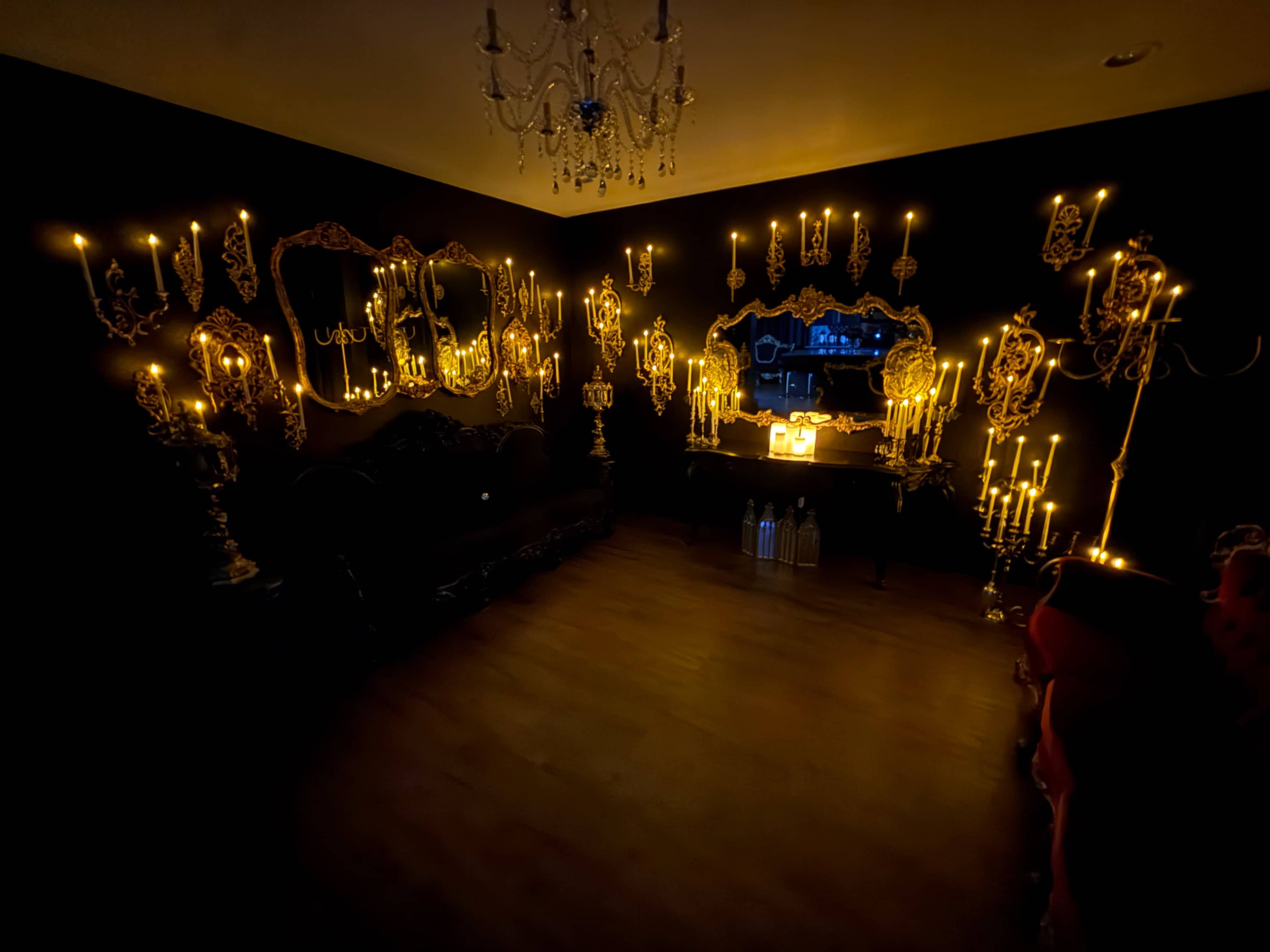 A dimly lit room featuring ornate mirrors and multiple candle holders that illuminate the dark walls.