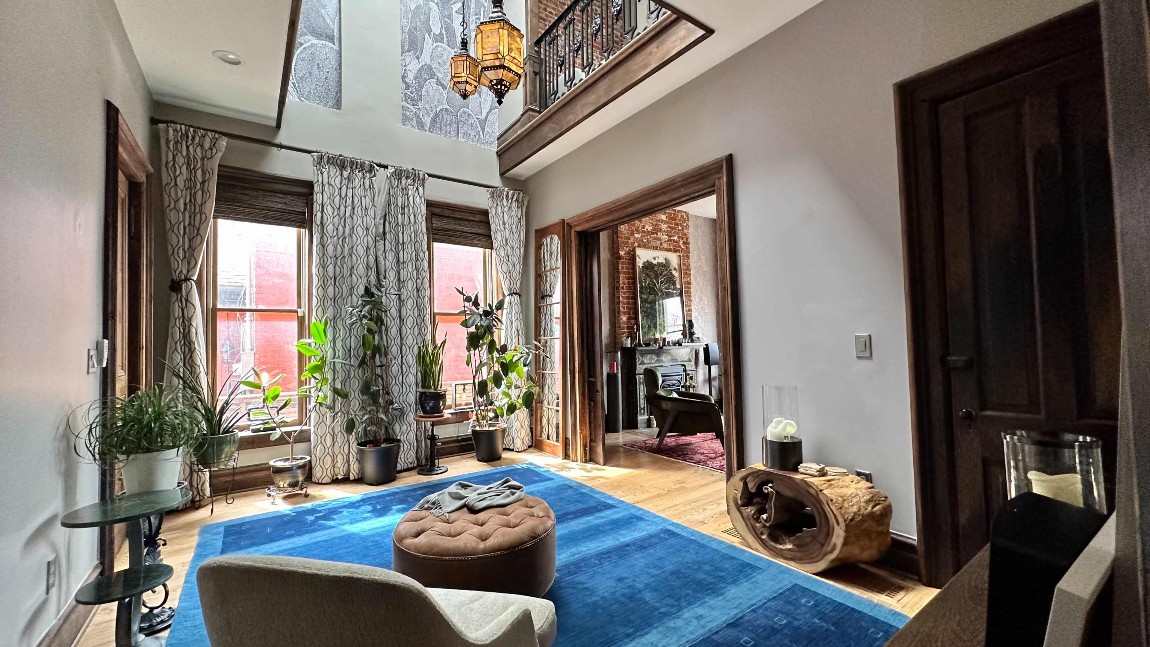 A stylish living room featuring a plush blue rug, a brown tufted ottoman, potted plants near large windows, and an upper balcony with decorative lanterns.
