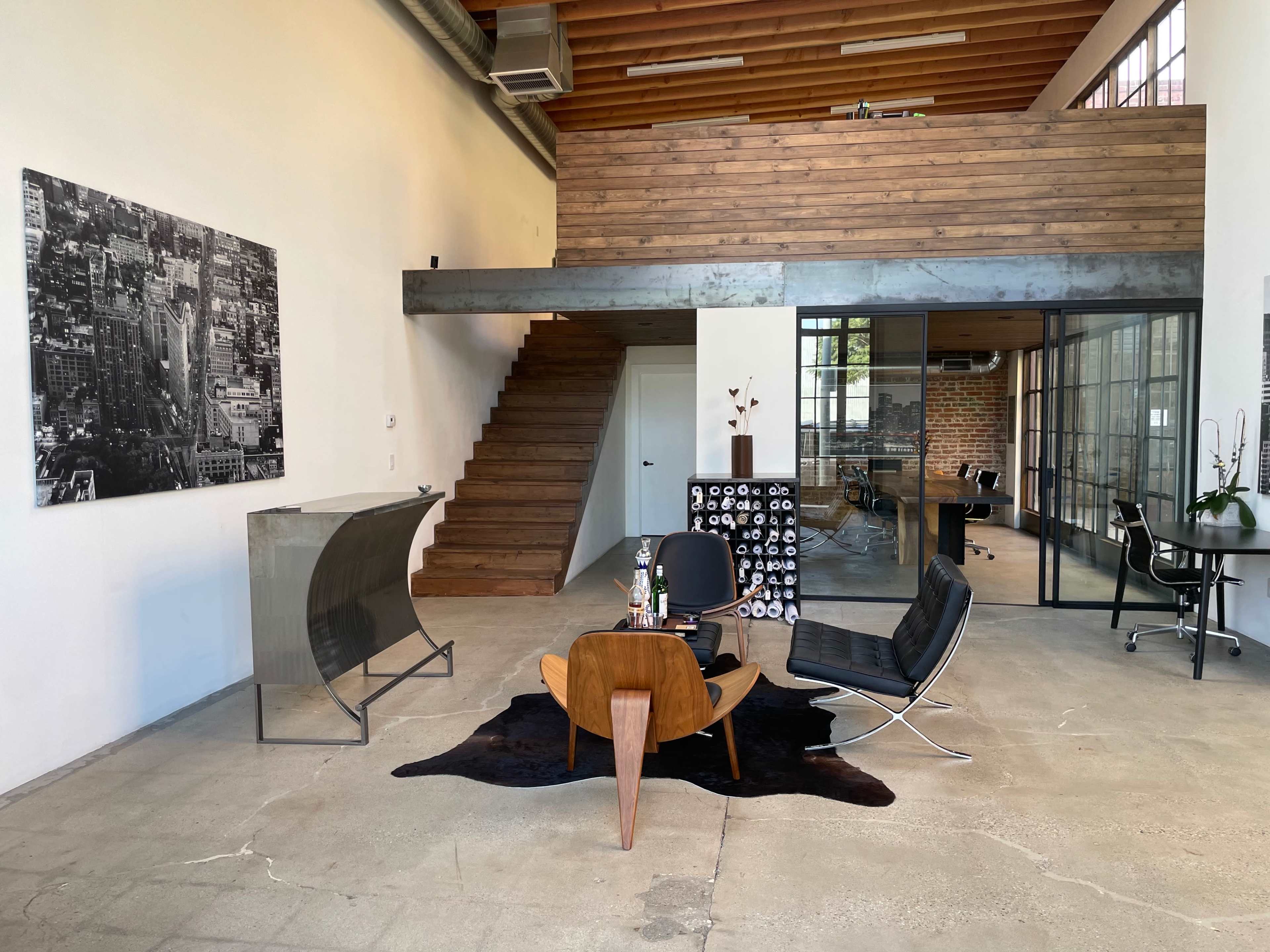 The image shows a spacious, modern office interior featuring a wooden staircase, a seating area with a coffee table and chairs, and a glass-walled meeting room.