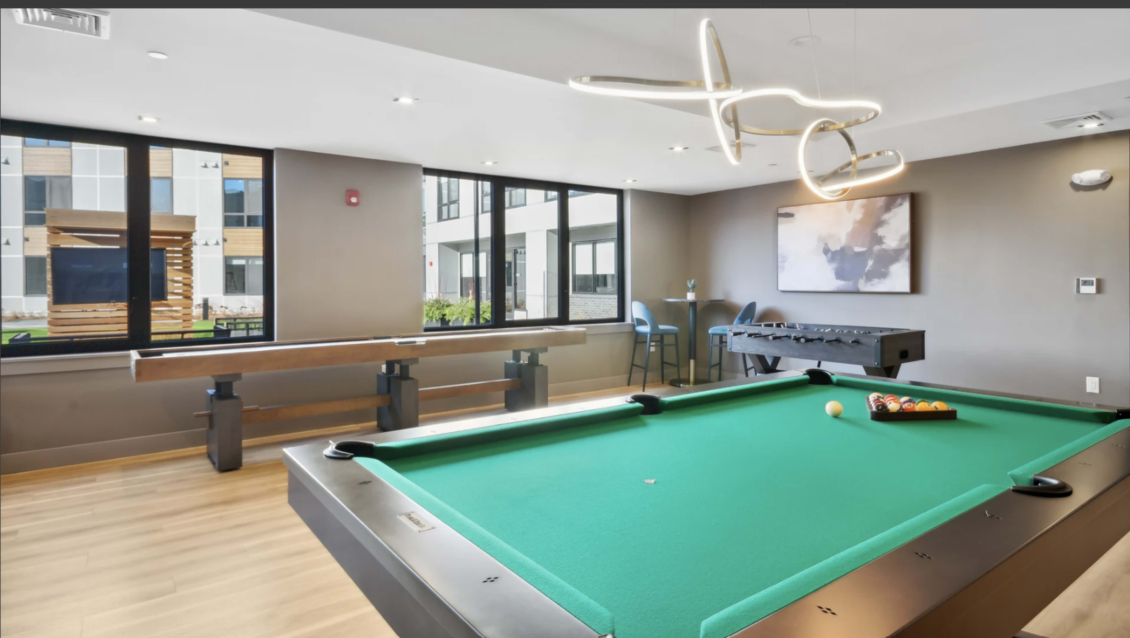 The image features a brightly lit room with a green billiards table, a foosball table, and large windows overlooking an outdoor area.