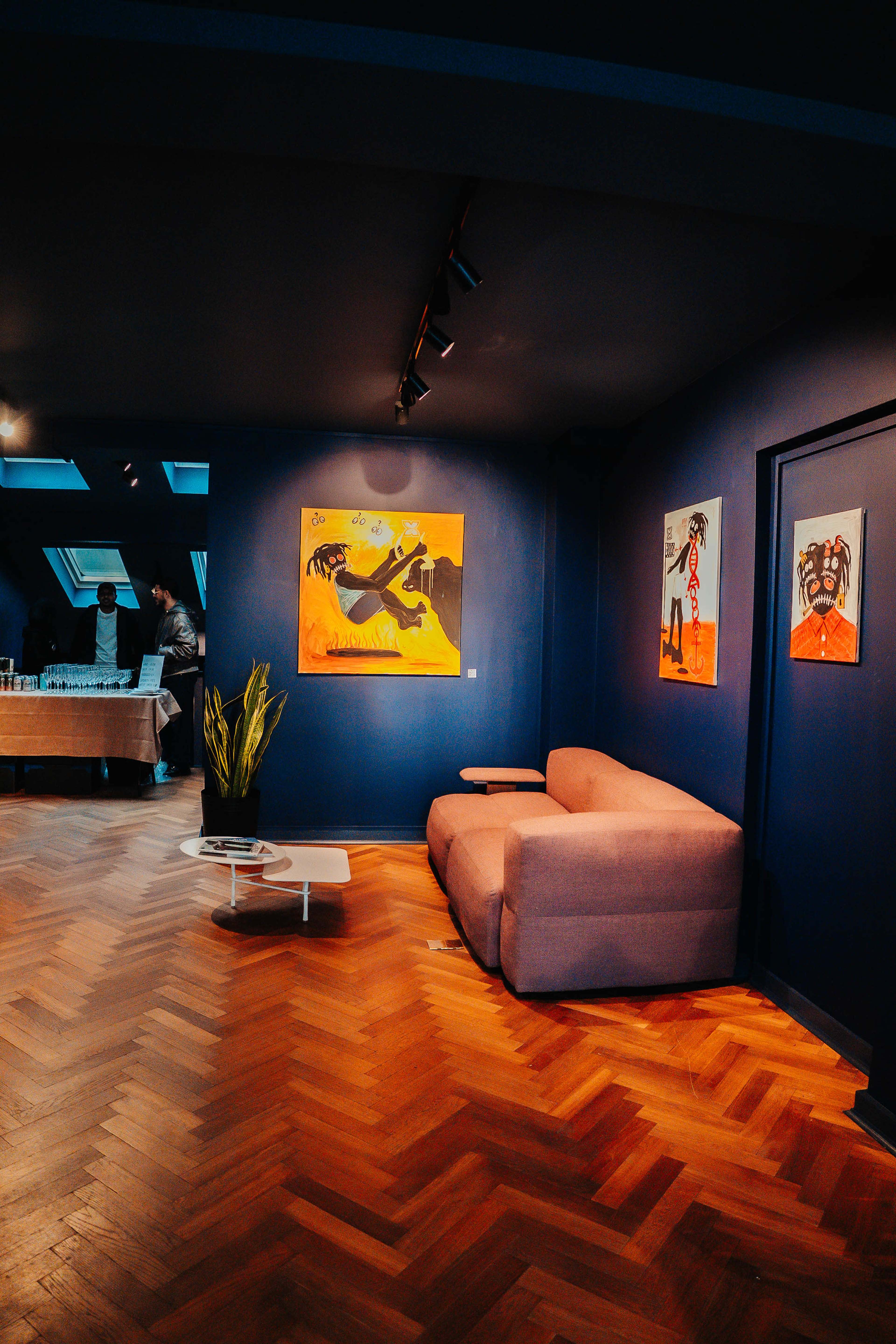A cozy room featuring a low sofa, dark blue walls adorned with colorful paintings, and a wooden herringbone floor.