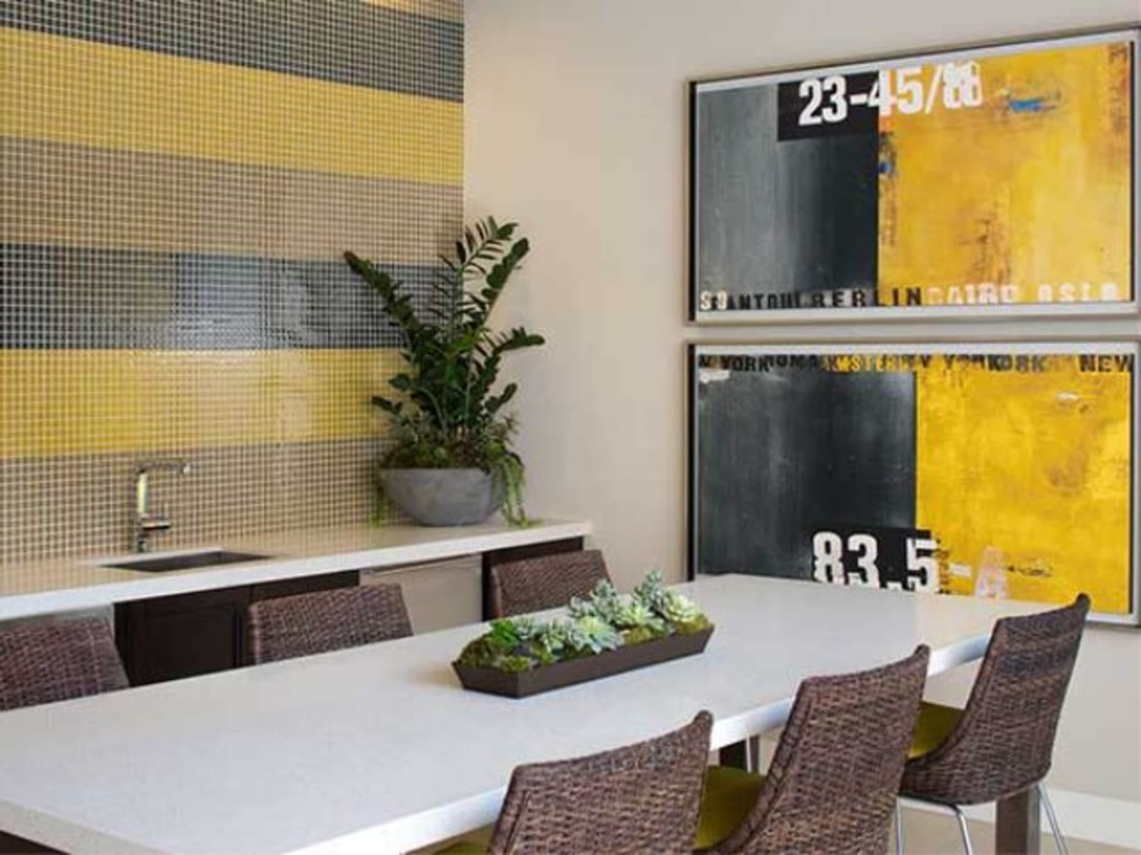 A modern dining area features a long table surrounded by woven chairs, with a mosaic wall and abstract art on display.