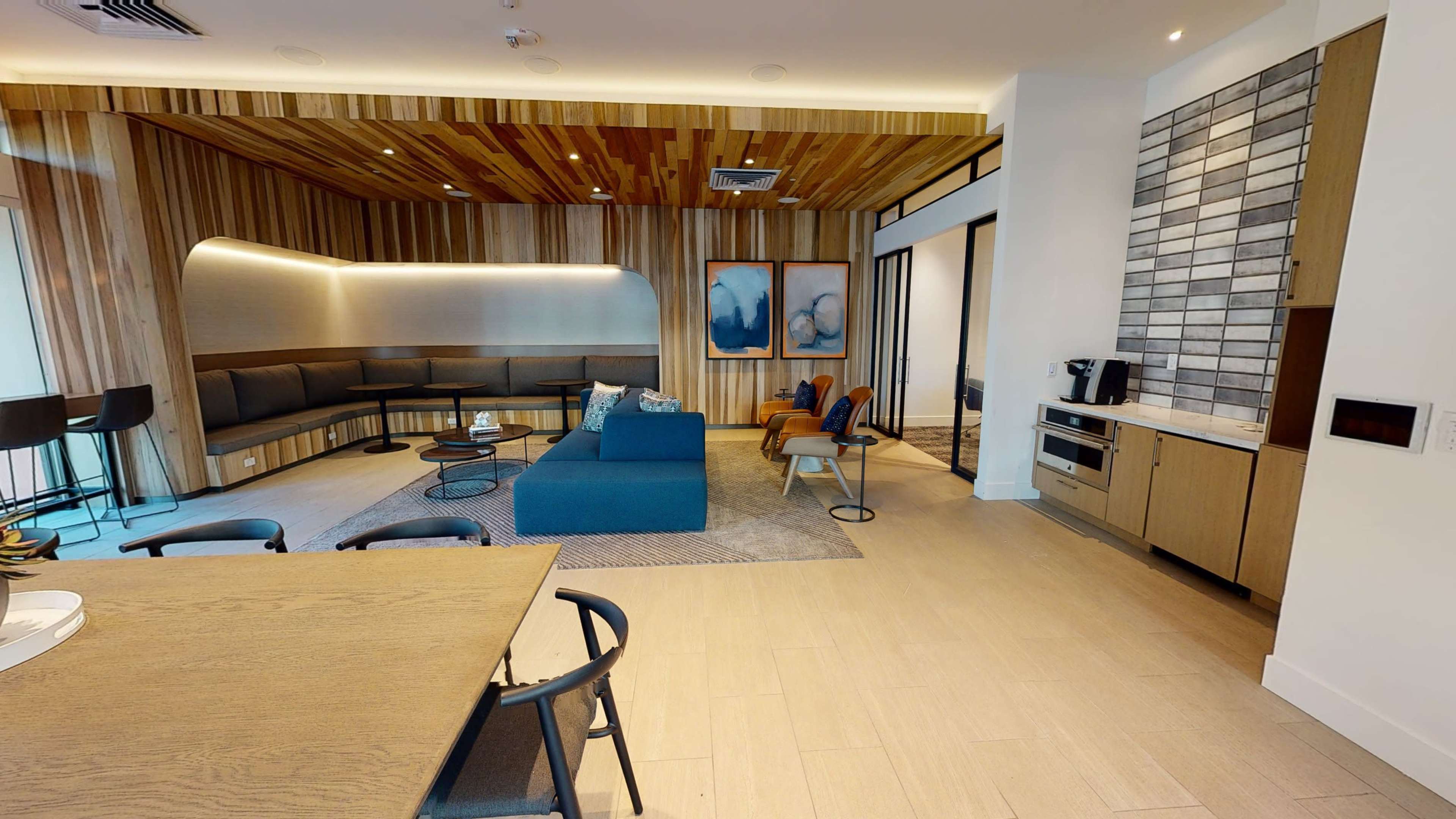 A modern communal lounge area with wooden paneling, a blue seating arrangement, a dining table, and a kitchenette in the background.