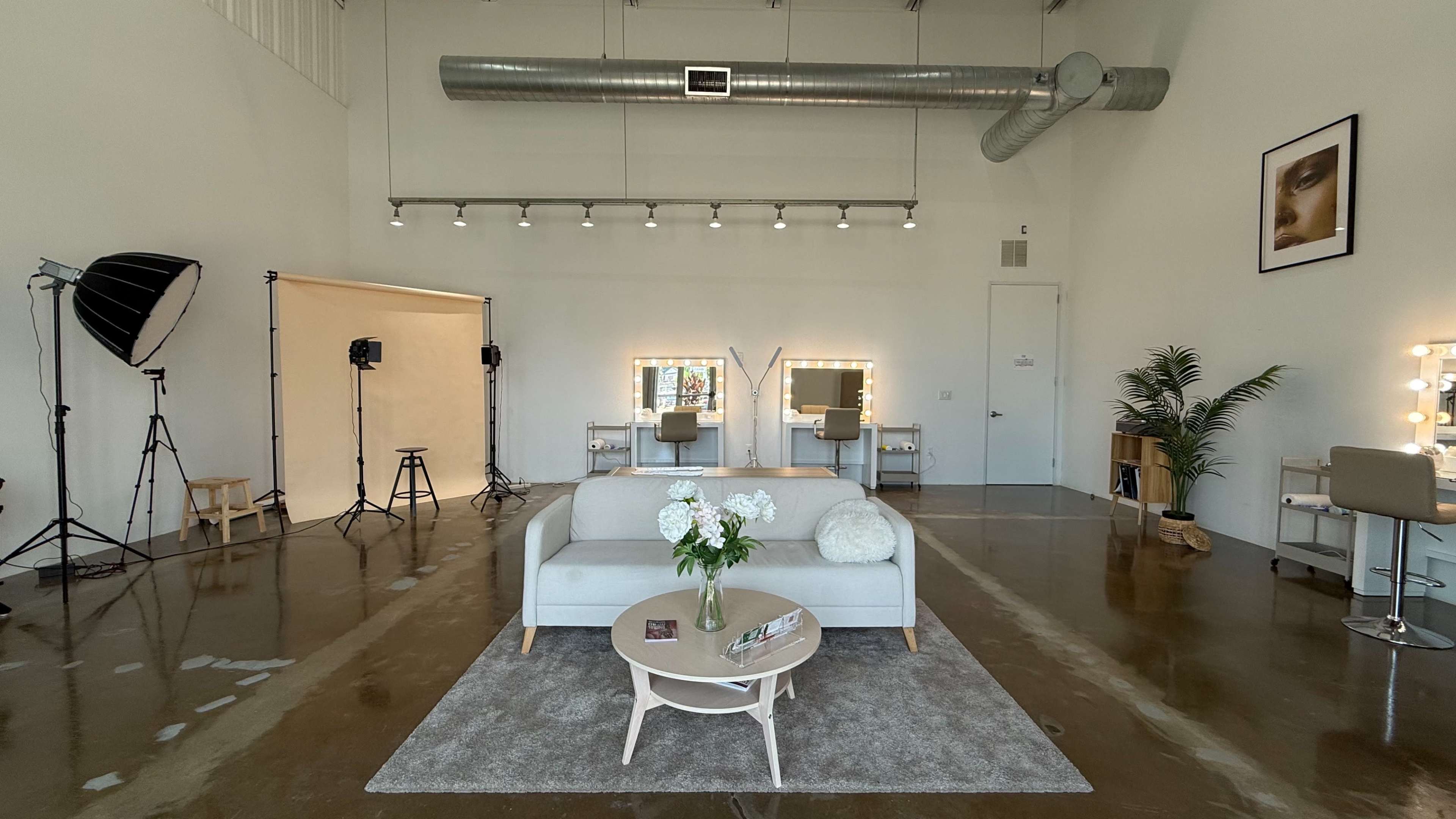 The image shows a spacious, minimalist studio with a light-colored sofa, a round coffee table, and multiple lighting setups arranged around the space.