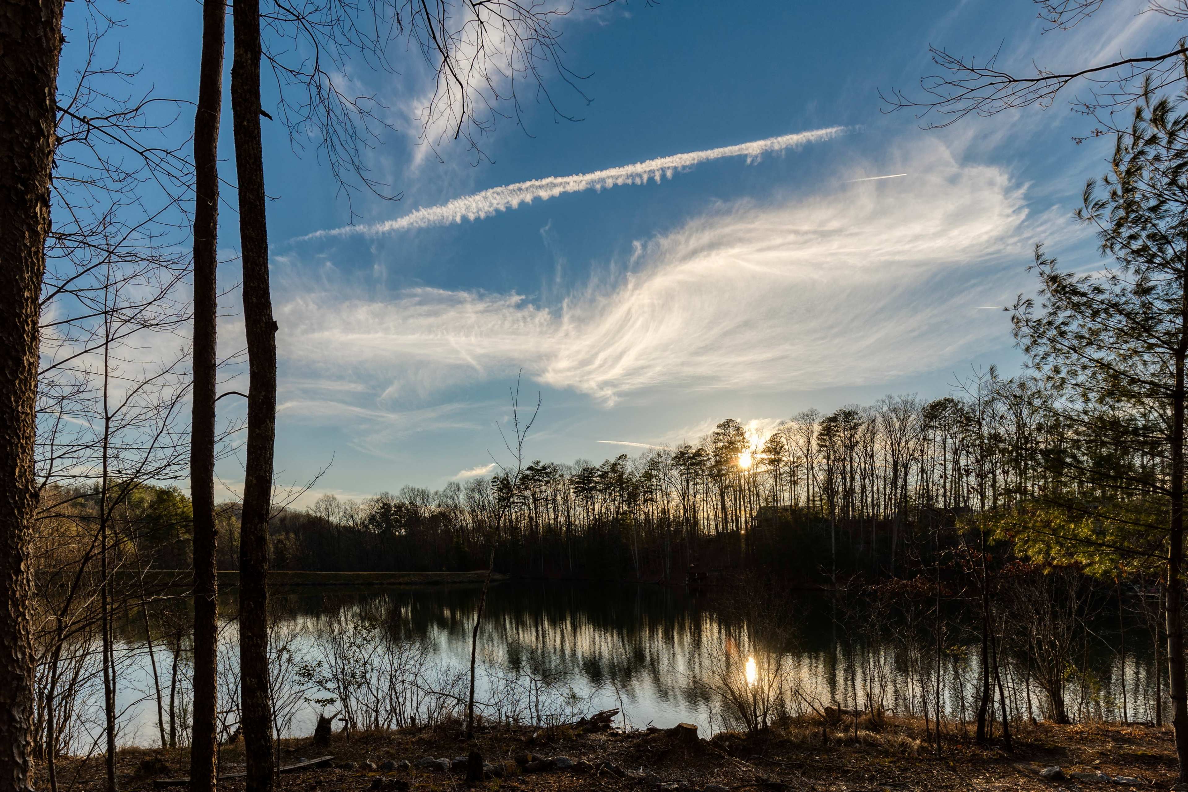 A calm lake surrounded by trees under a sky filled with wispy clouds and a setting sun.