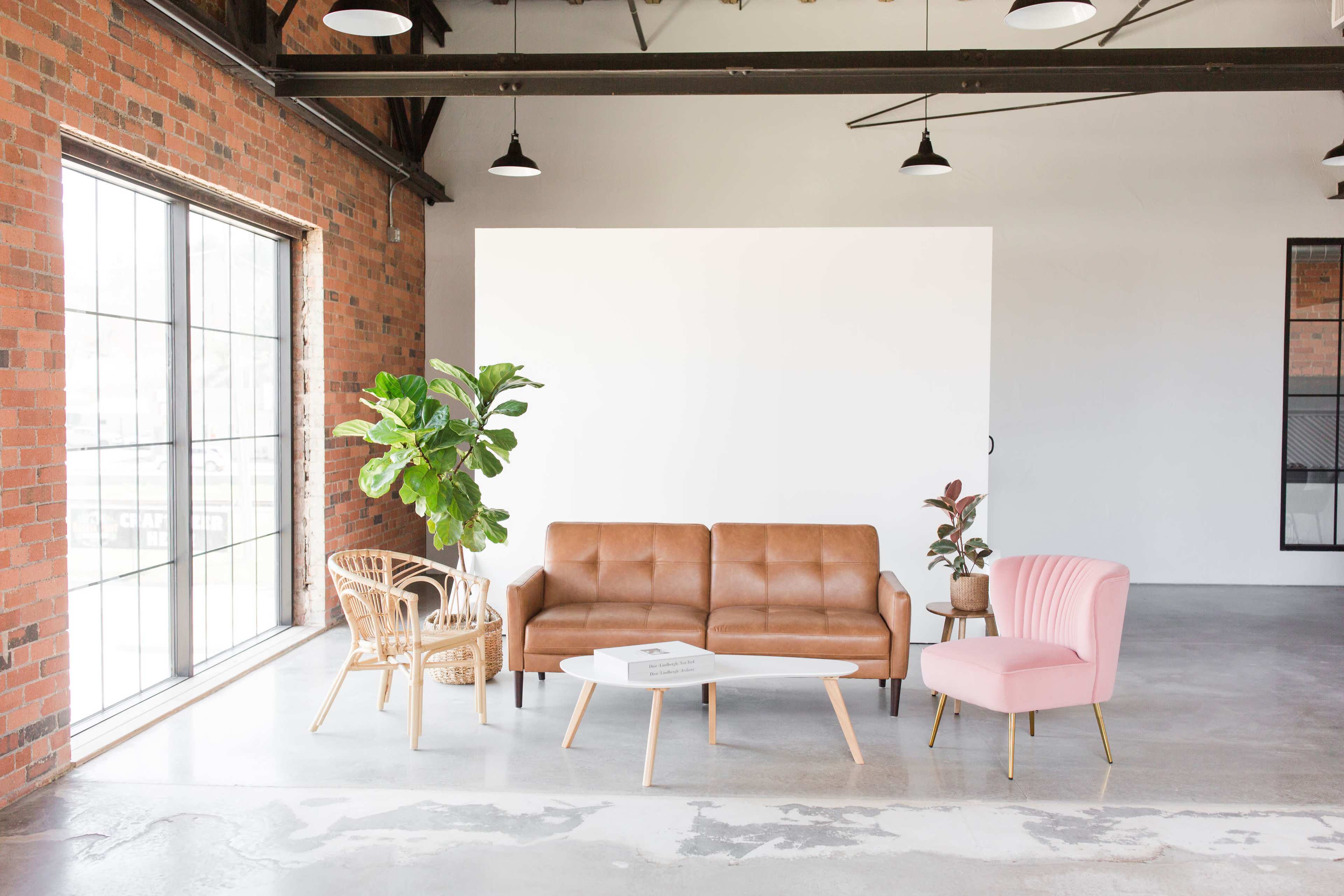 A modern interior space featuring a brown leather sofa, a pink armchair, a wooden chair, and a coffee table, complemented by greenery and an industrial-style brick wall.