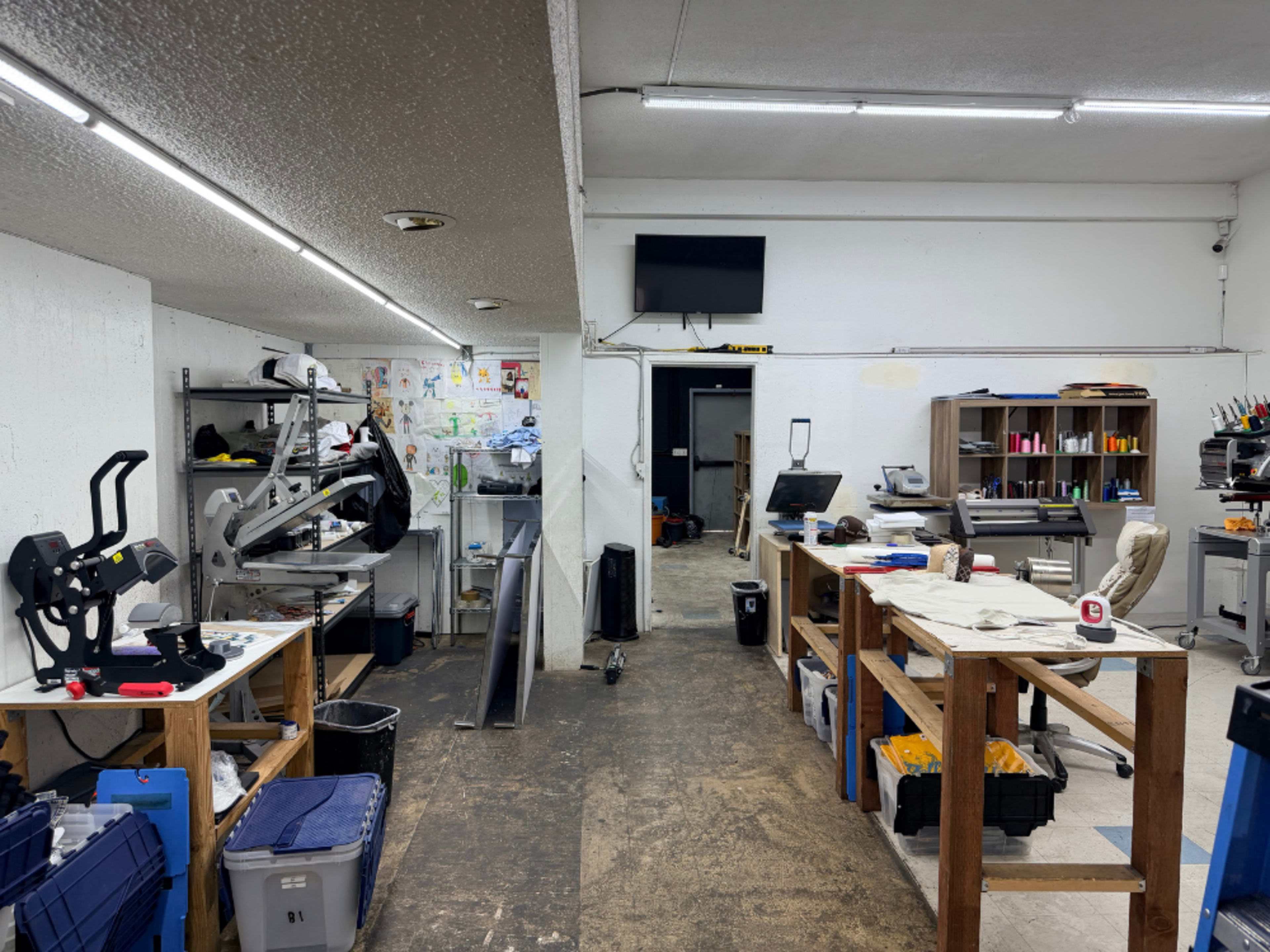 The image shows a workshop space with wooden tables, tools, and equipment arranged on both sides, featuring a TV mounted on the wall and an office-like area in the background.
