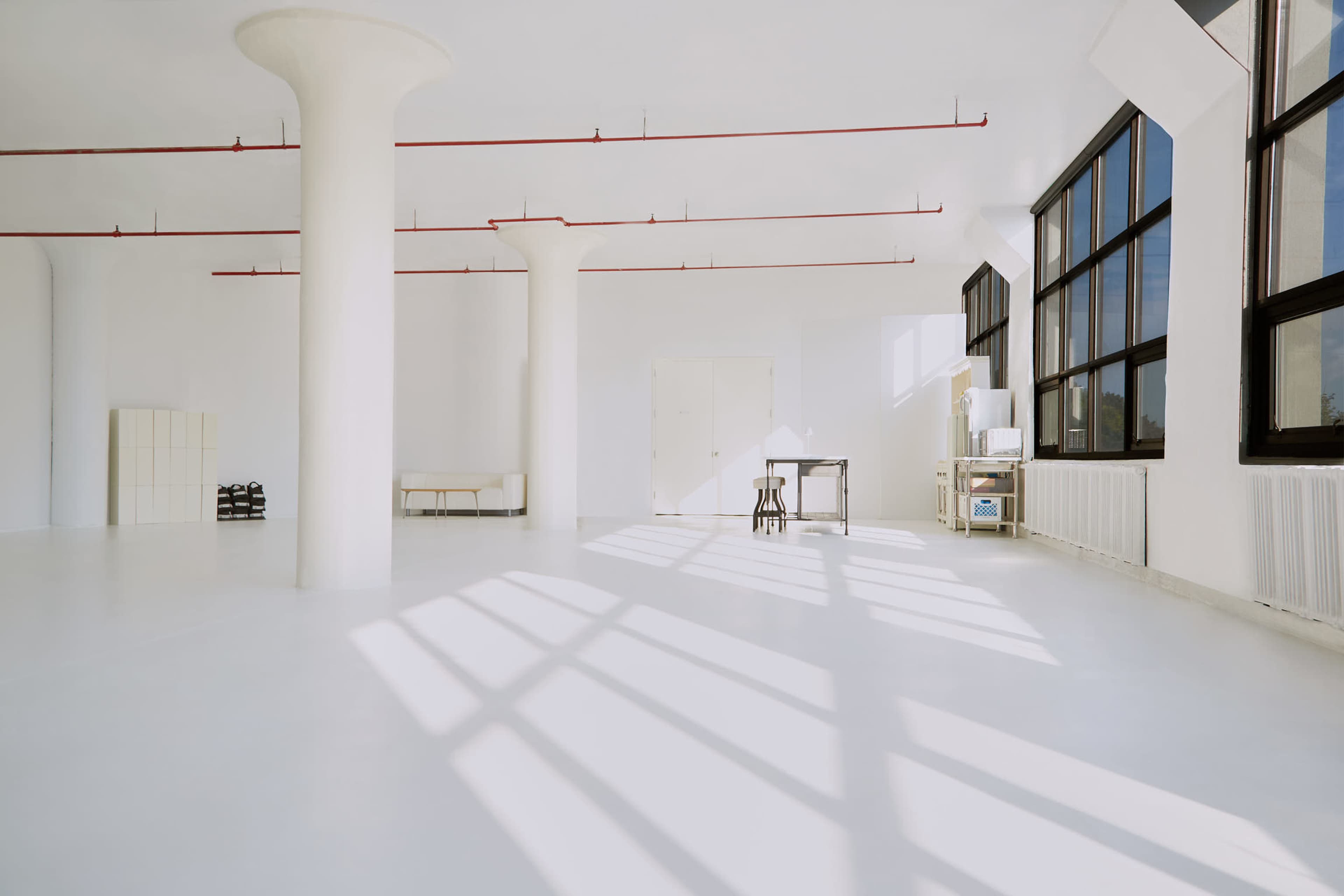 A spacious, bright room with white walls and floors, featuring large windows, a minimalistic table, and a few pillars.