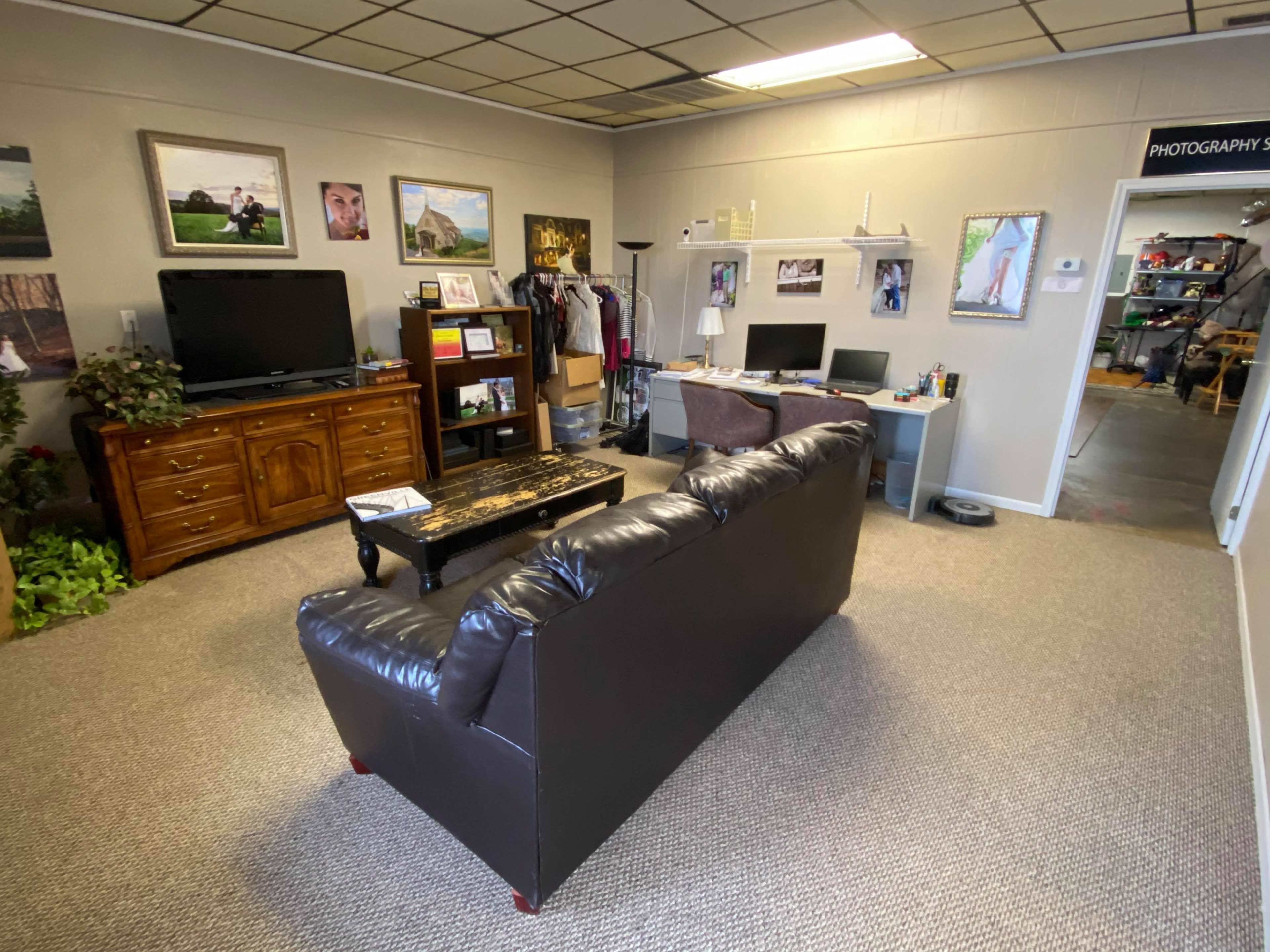 The image shows a small office space featuring a dark leather sofa, a wooden television cabinet, a desk with a computer, and framed photographs on the walls.