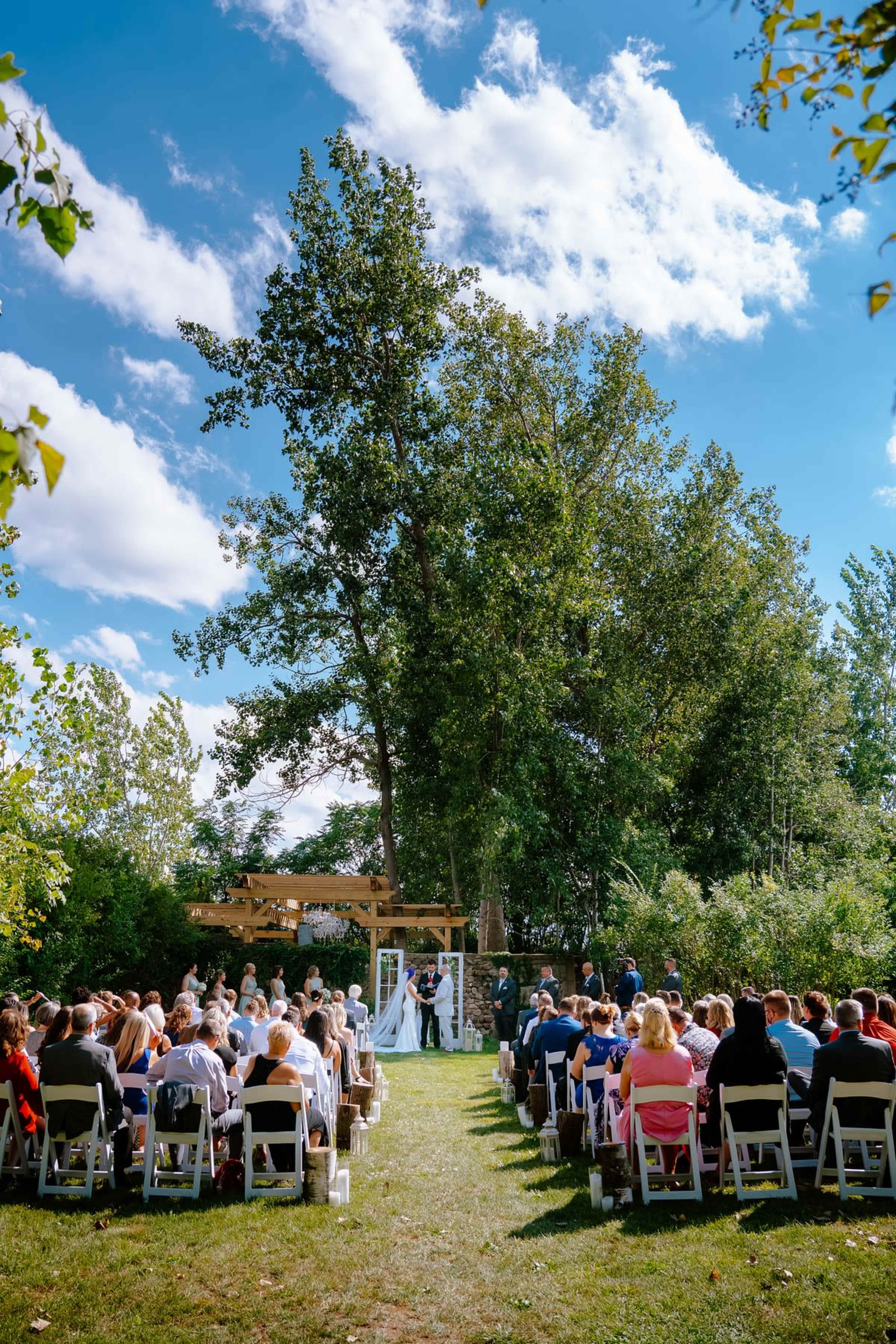 A wedding ceremony takes place outdoors under a wooden arch, surrounded by trees and seated guests.