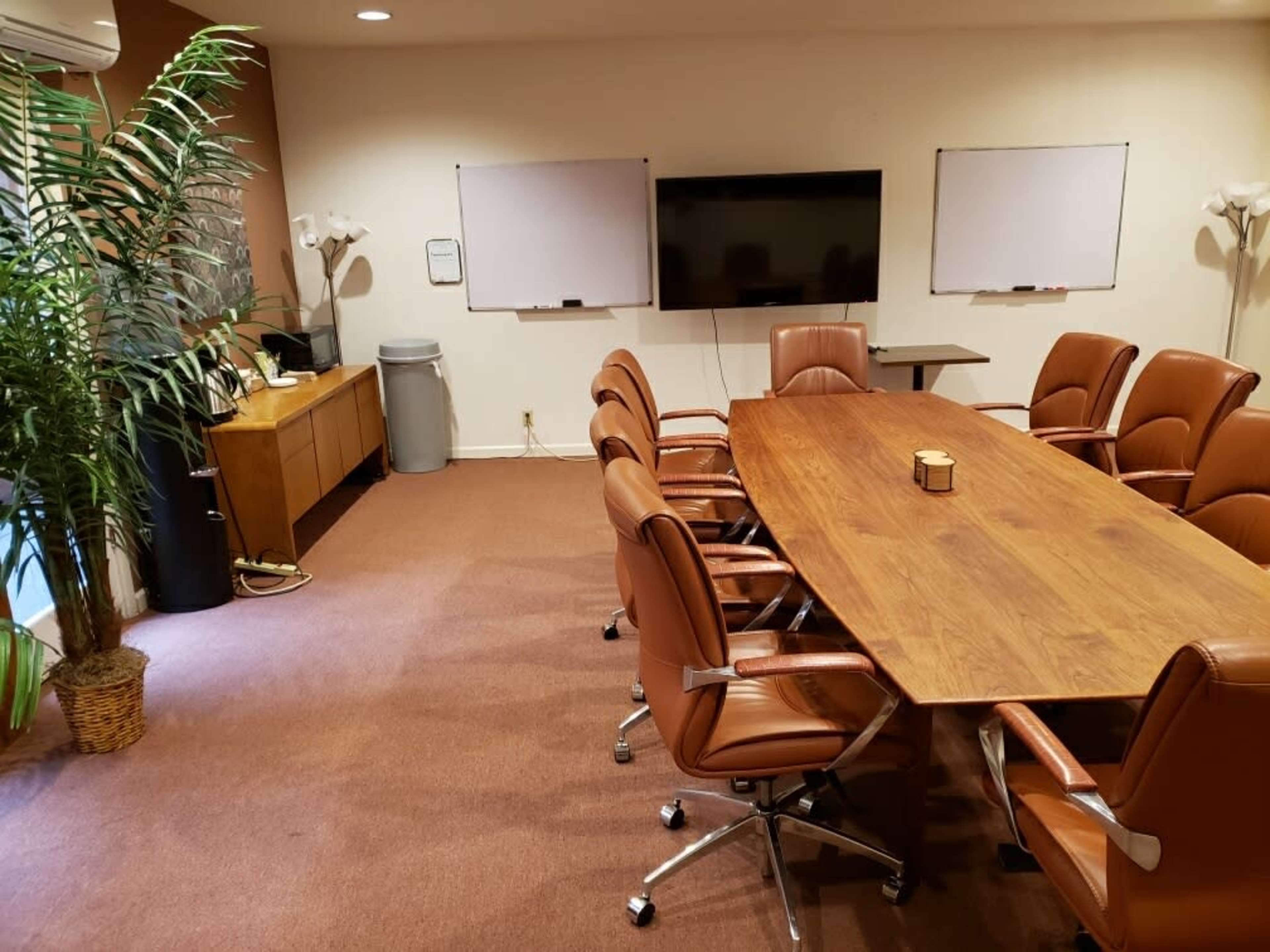A conference room features a large wooden table surrounded by brown leather chairs, with a wall-mounted screen and whiteboards in the background.