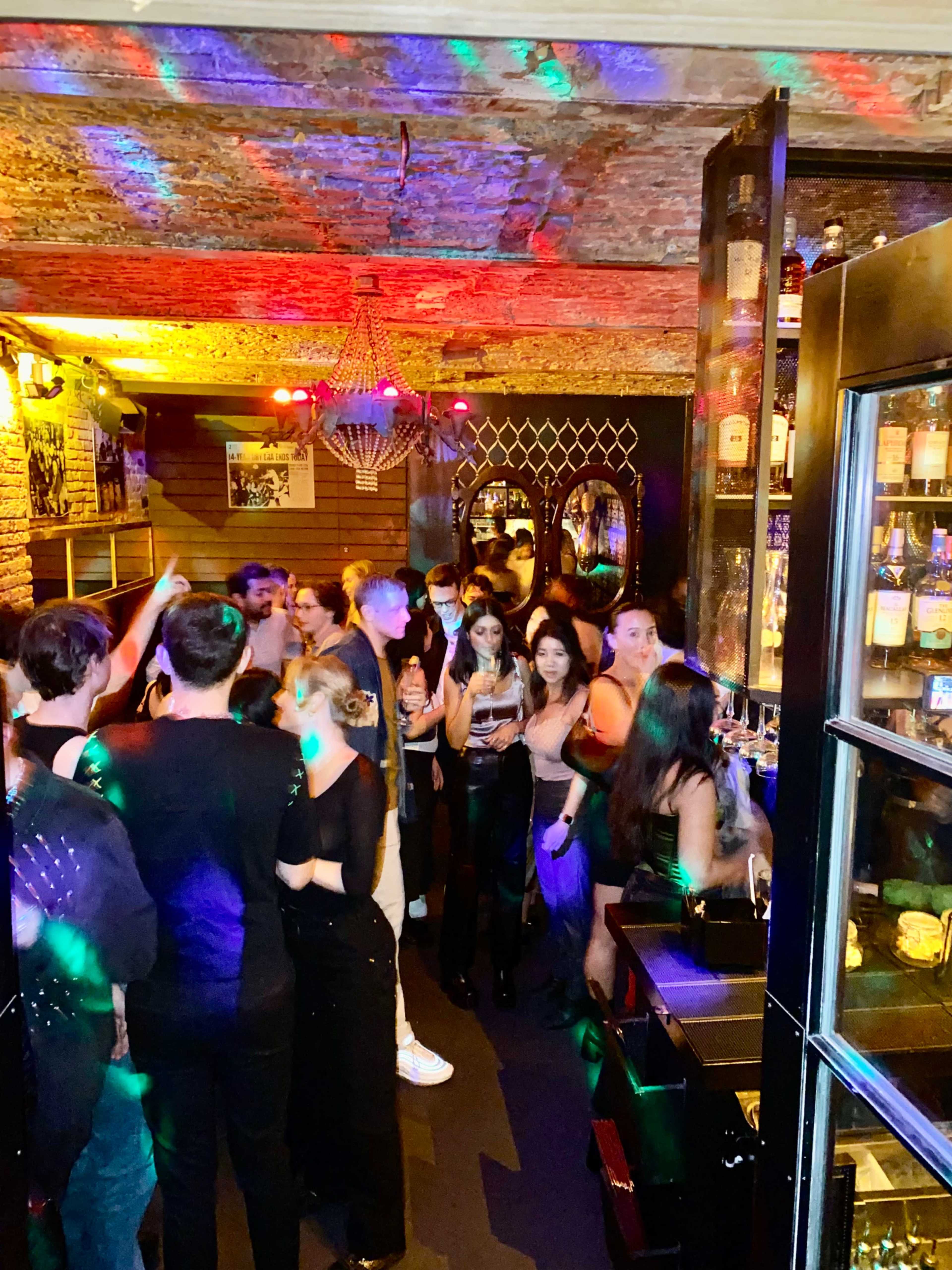 A crowd of people socializes in a dimly lit bar with colorful lighting and a display of liquor bottles.
